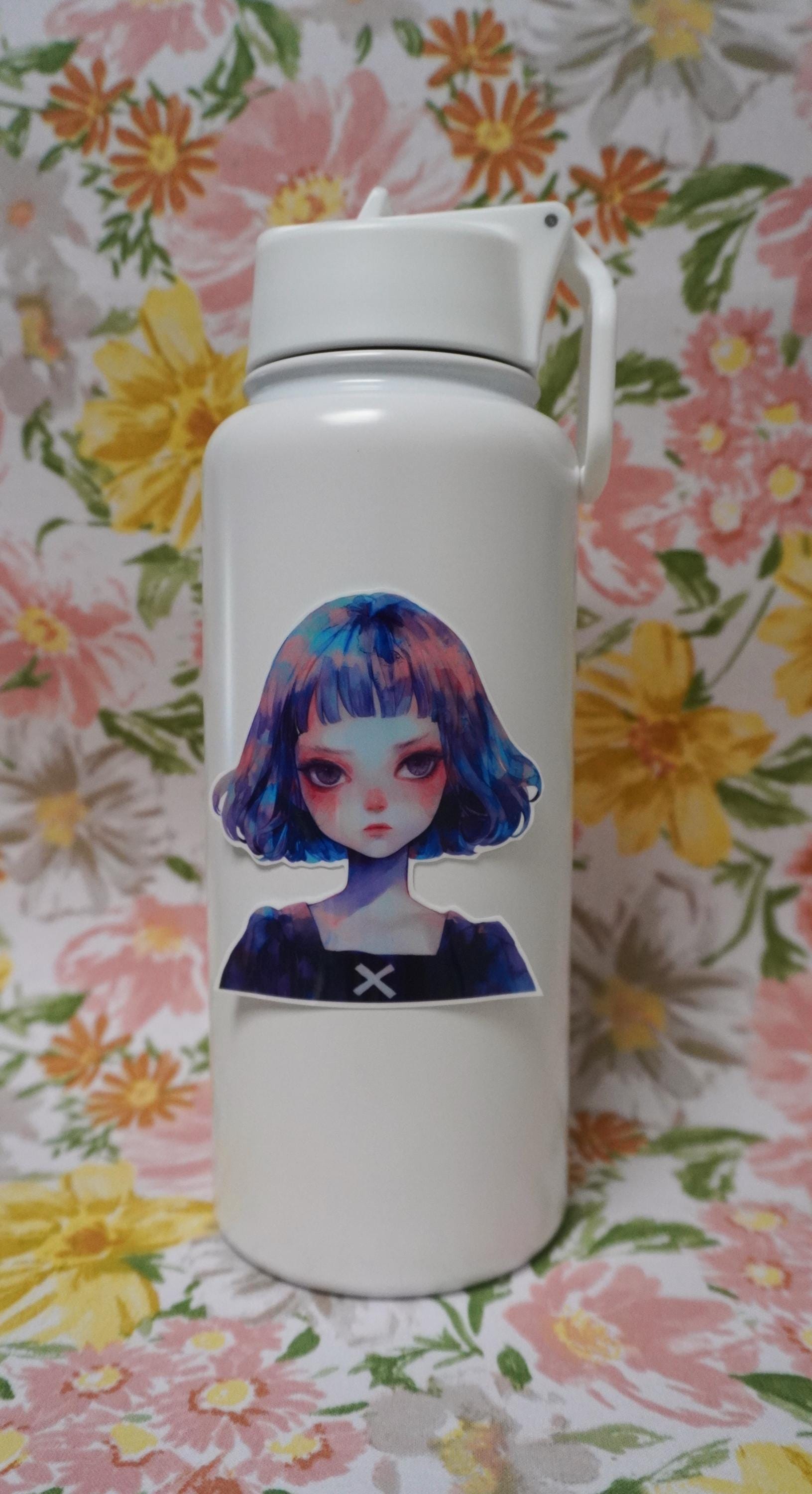 A white water bottle decorated with a sticker of a girl with blue and purple hair. The background is a floral fabric with large yellow, pink, and orange flowers, along with green leaves.