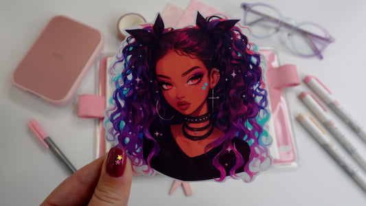 A hand holding a sticker of a girl with curly purple and blue hair, wearing chokers and star face decals. Background includes a pink notebook, markers, washi tape, paper, a compact printer, and glasses. Nails are dark pink with star accents.