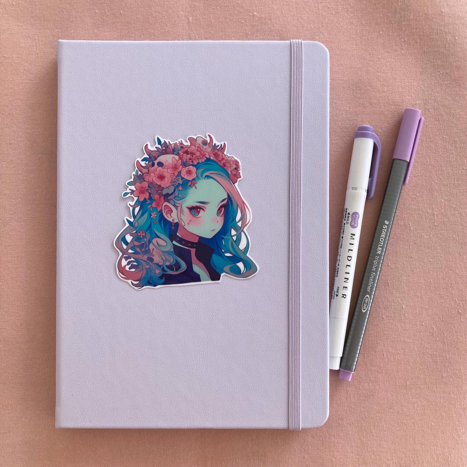 A sticker of a girl with blue hair and a floral crown on a light lavender notebook. Beside the notebook are a purple marker and a gray pen, all set on a soft pink fabric background.