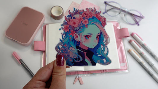 A hand holds a sticker of a girl with long blue hair and a floral crown, wearing a black choker. The background includes a pink notebook, markers, washi tape, pink paper, a compact printer, and glasses. Nails are dark pink with a silver star accent.