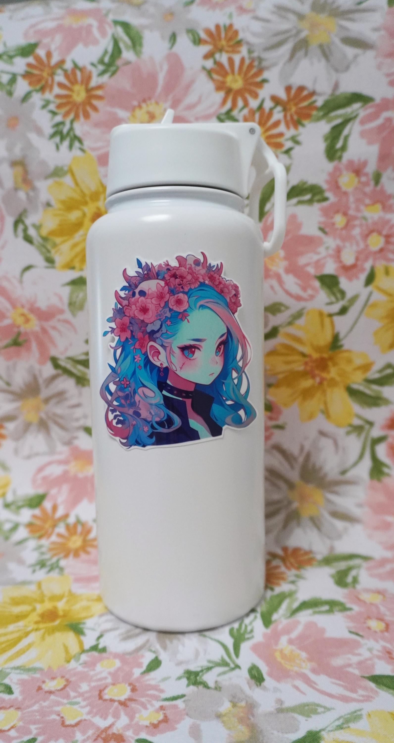 A white water bottle with a sticker of a girl with blue hair, a floral crown, and a black choker. The background is a floral fabric with large pink, yellow, and orange flowers, and green leaves.
