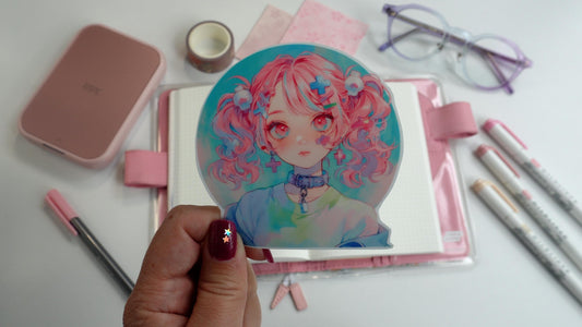 A hand holds a sticker of a girl with pink hair in pigtails, wearing a collar and pastel clothing. The background includes a pink notebook, markers, washi tape, pink paper, a compact printer, and glasses. Nails are dark pink with star accents.