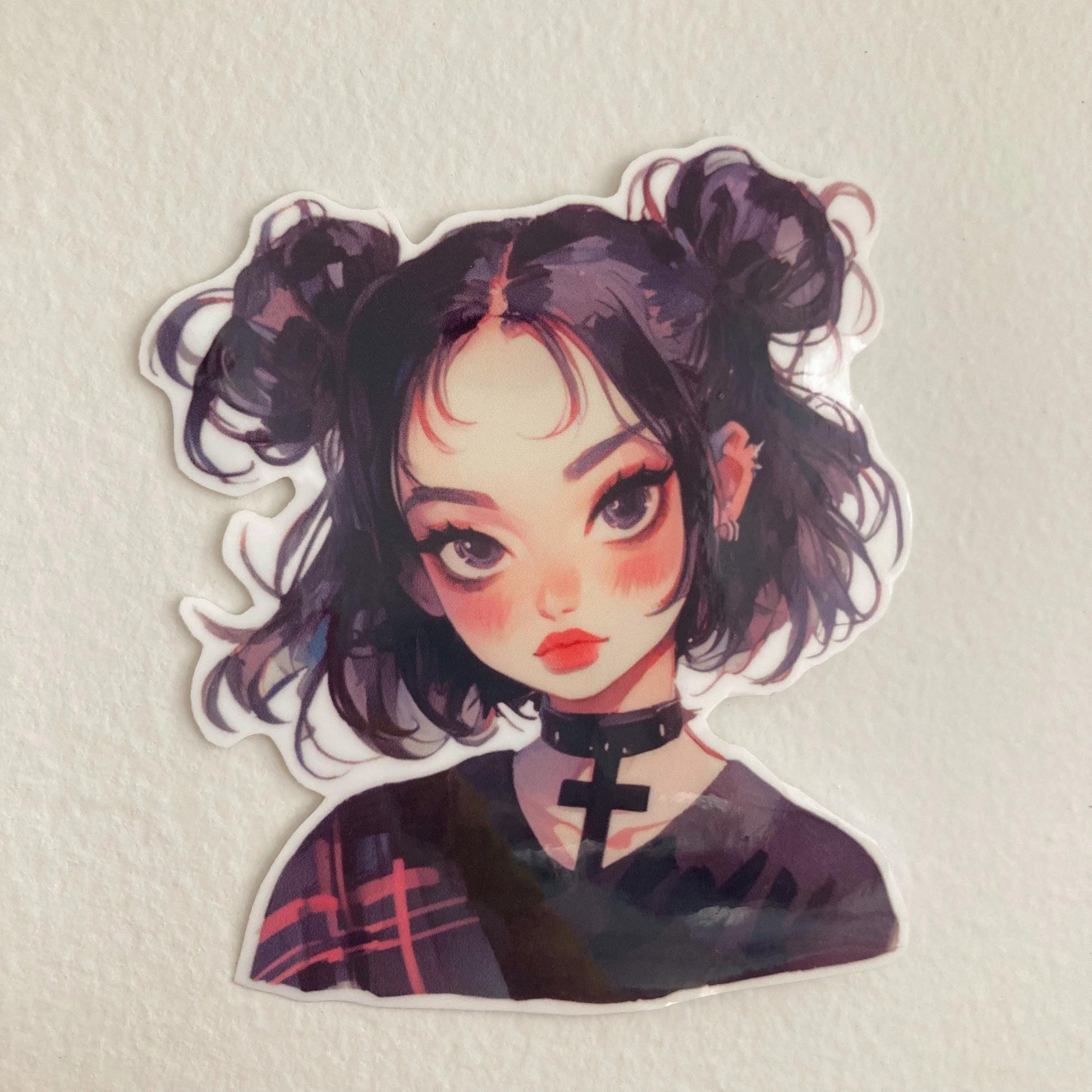 A sticker of a girl with black hair in buns, wearing a choker and a dark shirt with plaid accents, placed on a plain light-colored background.