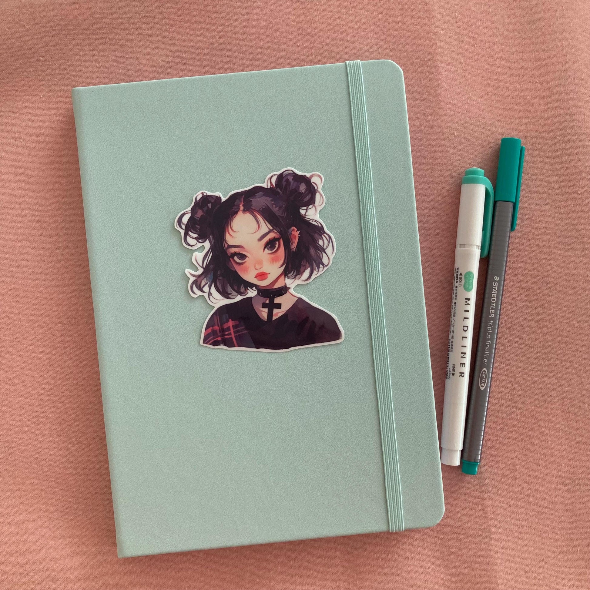 A sticker of a girl with black hair in buns and a choker is placed on a light teal notebook. Beside the notebook are a green marker and a gray pen, all set on a soft pink fabric background.
