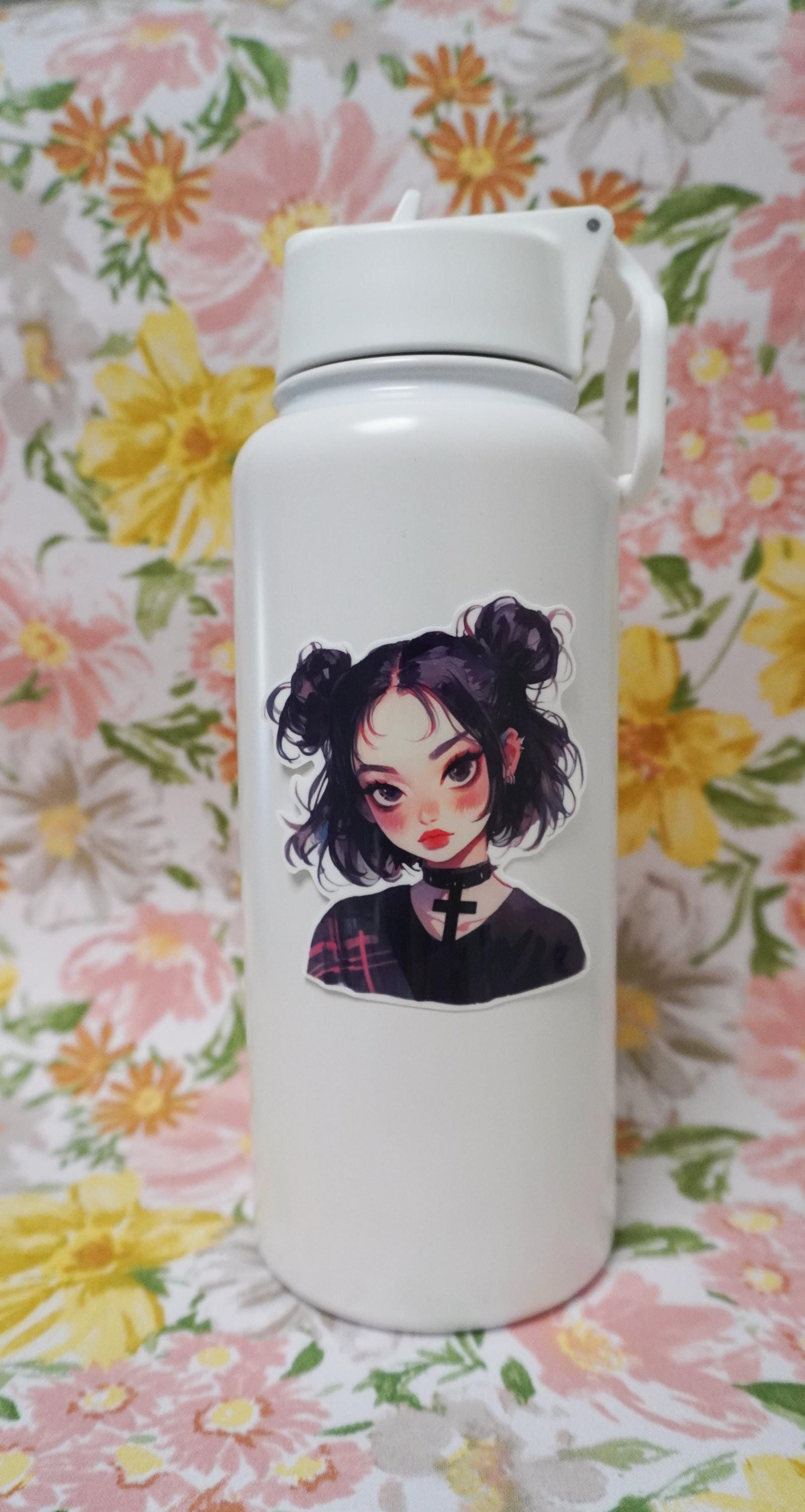 A white water bottle with a sticker of a girl with black hair in buns, wearing a choker and a dark shirt. The background is a floral fabric with pink, yellow, and orange flowers and green leaves.