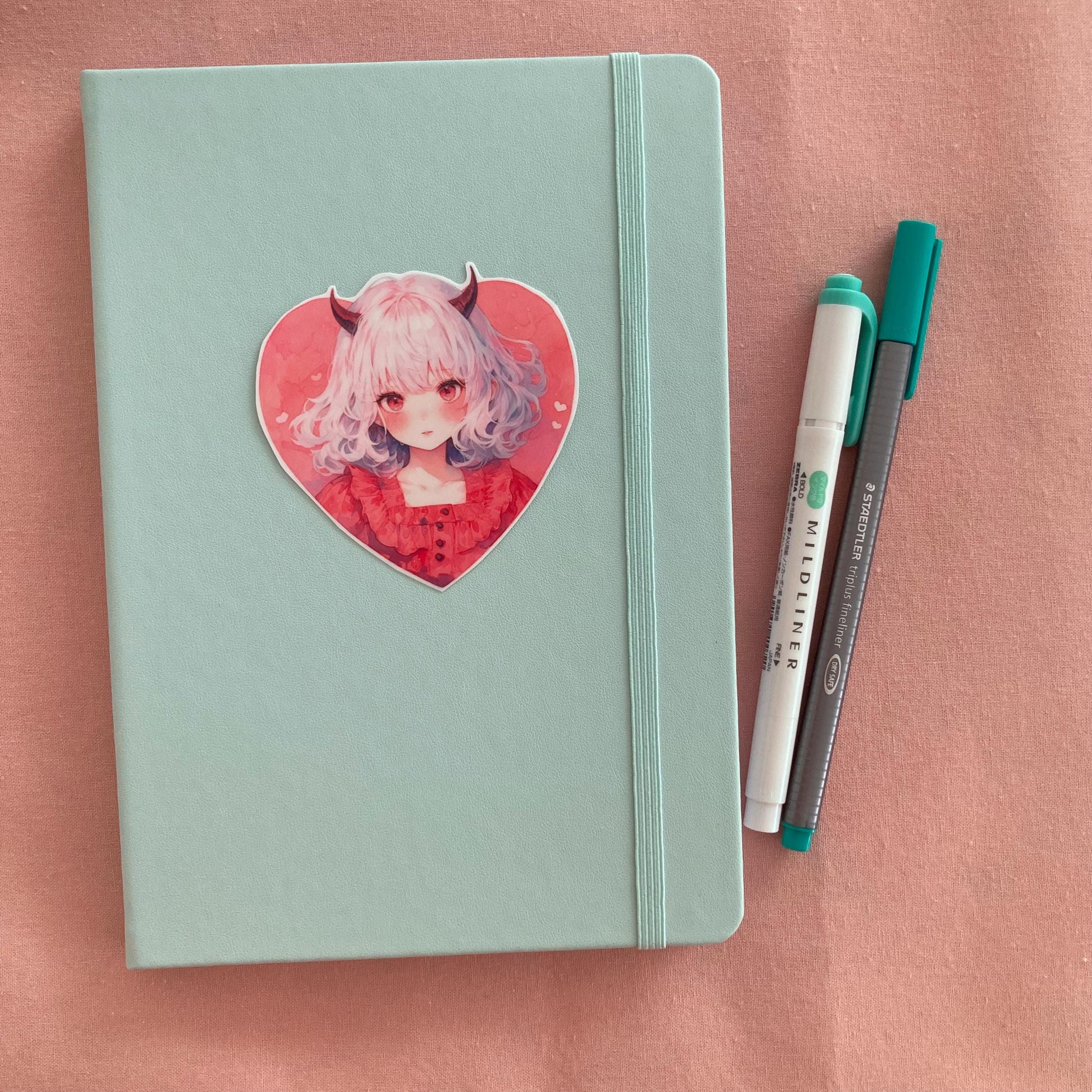 A heart-shaped sticker of a girl with white hair, red horns, and a red dress is placed on a light teal notebook. Beside the notebook are a green marker and a gray pen, all on a soft pink fabric background.