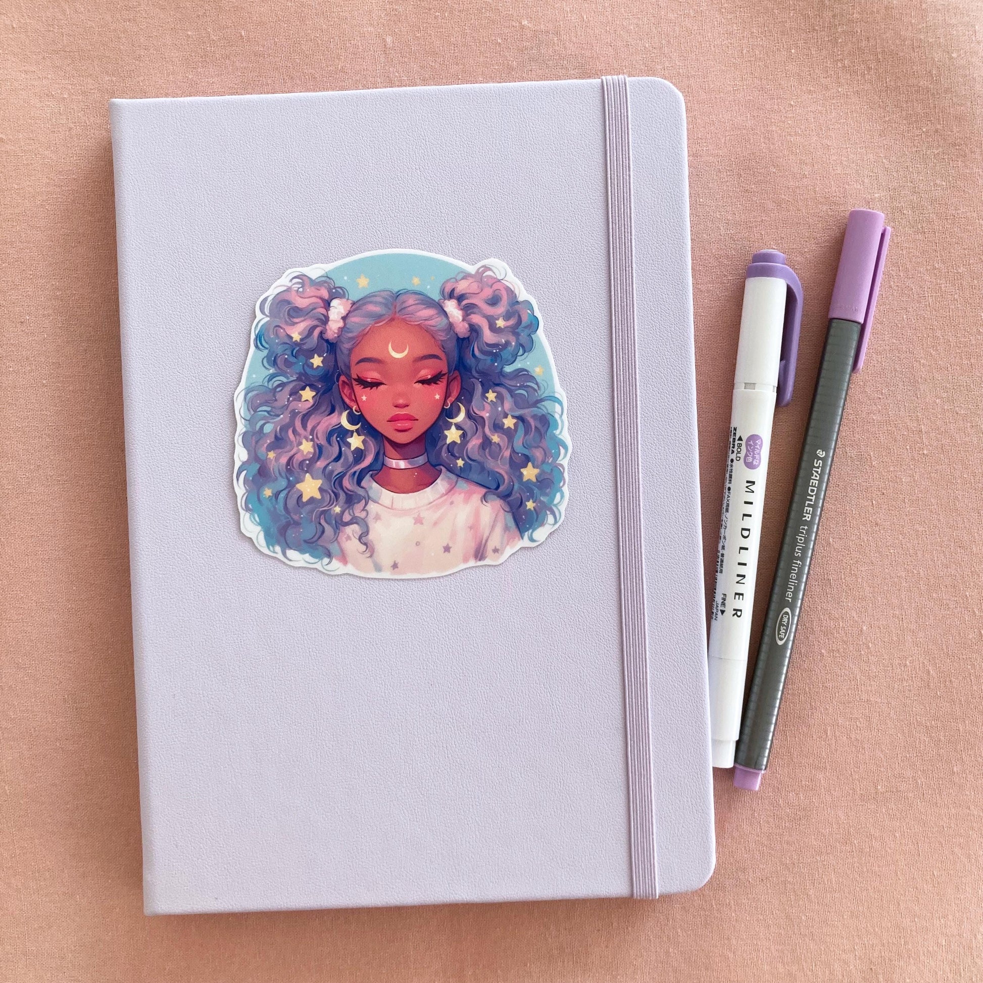 A pastel lavender notebook with an anime-style sticker of a girl with lavender and blue curly hair on its cover, next to a purple marker and gray pen on a pink fabric background.