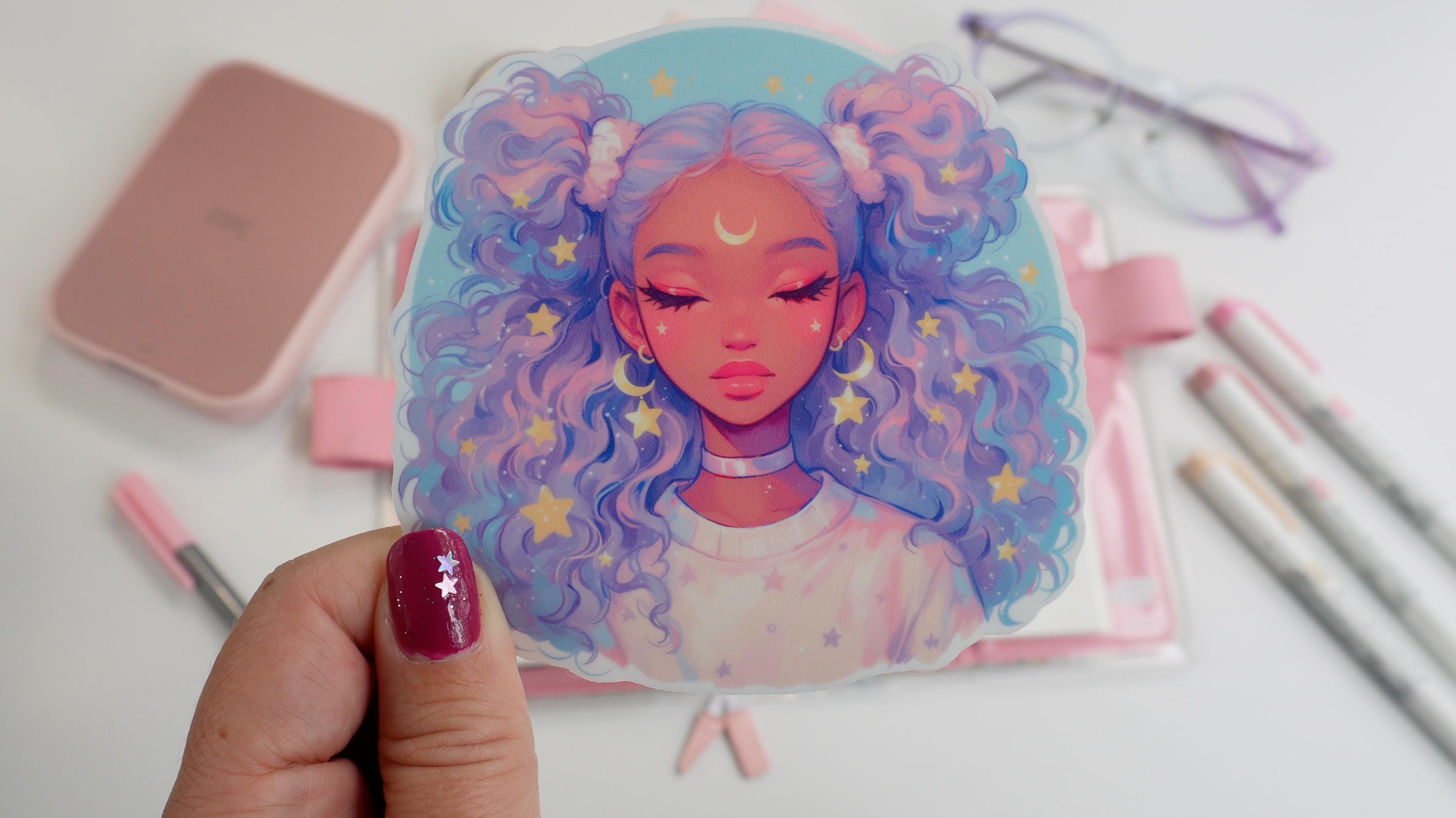 A hand holding a sticker of an anime-style girl with lavender and blue curly hair, closed eyes, and a crescent moon on her forehead. Background includes pink stationery, an open notebook, pastel markers, and glasses on a white surface.
