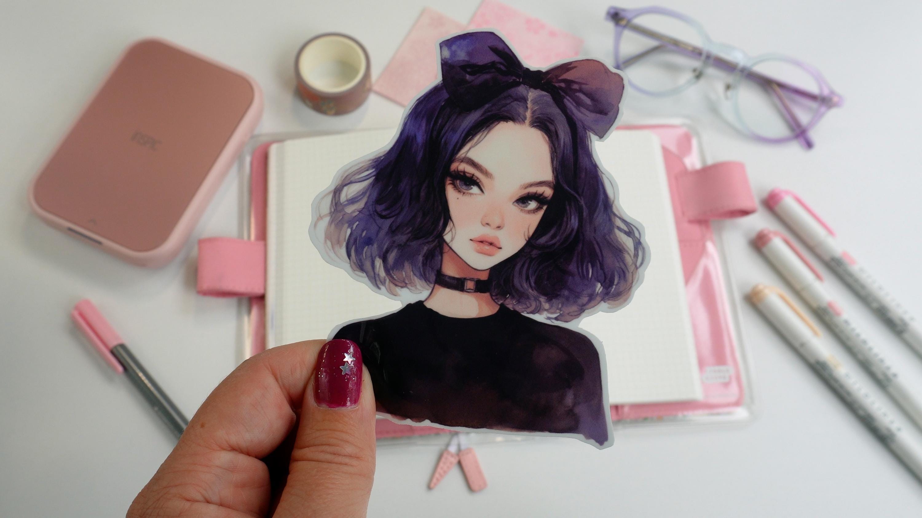 A hand holding a sticker of an anime-style character with short dark purple hair, a large bow, and a black choker. The background features pink stationery, an open notebook, pastel markers, and glasses on a white surface.