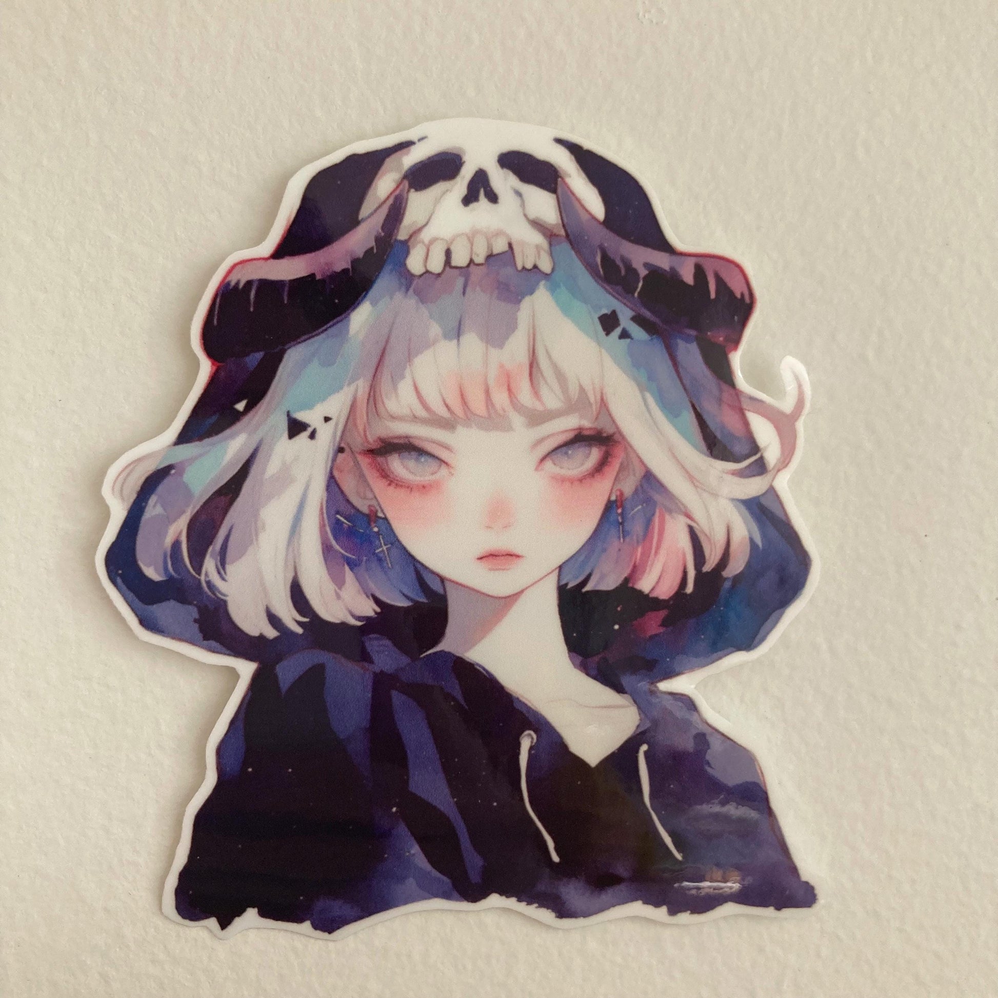 A sticker of an anime-style character with silver hair, wearing a dark hood with a skull design, placed on a plain white surface.