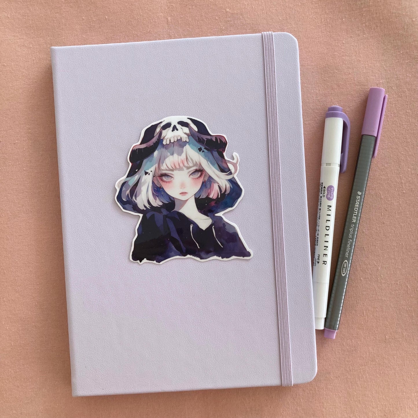 A pastel lavender notebook with a sticker of an anime-style character wearing a hood with a skull design, placed next to a purple marker and gray pen on a pink fabric background.