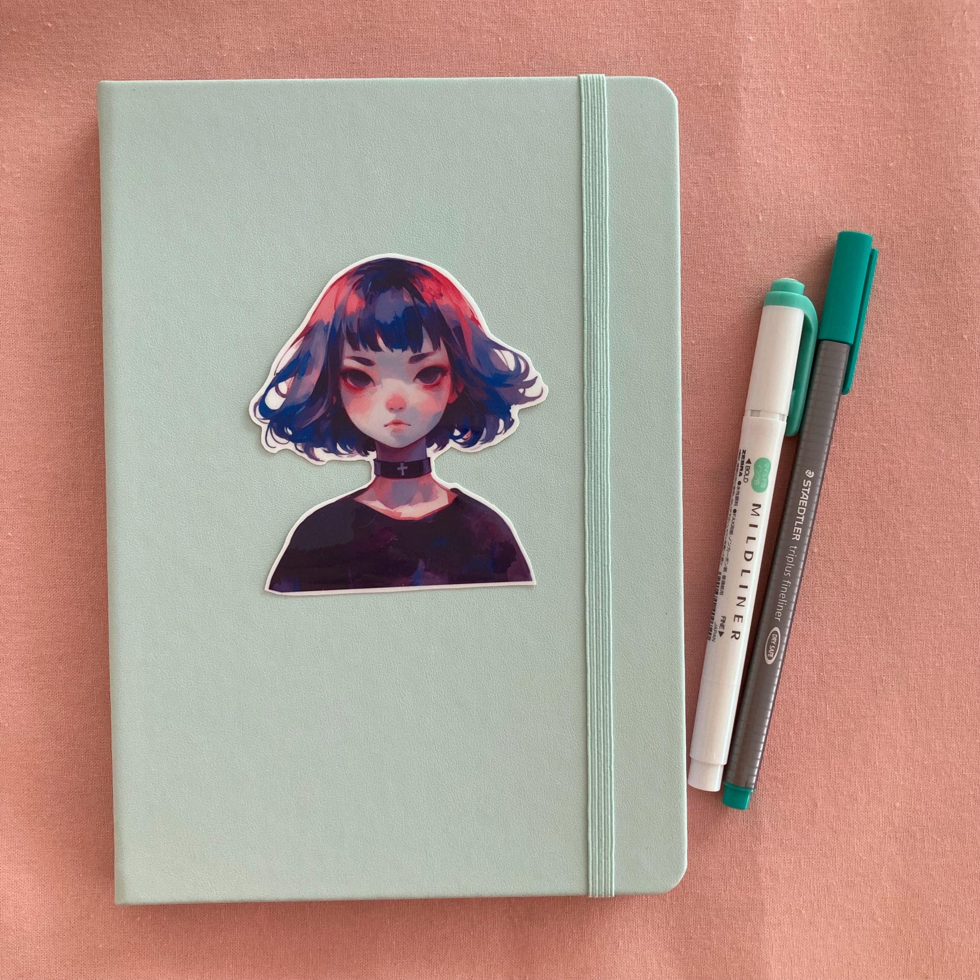Sticker of an anime-style character with blue, pink, and purple hair and a black choker with a cross, placed on a mint green notebook. Two pens, one green and one gray, are to the right. The background is a soft pink surface.