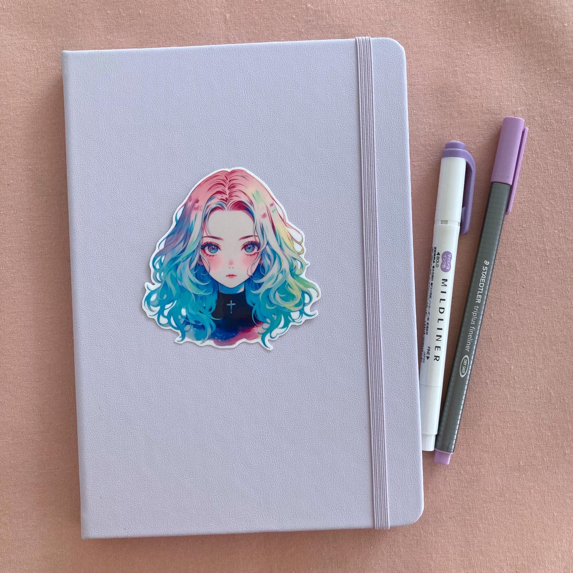 Lavender notebook with an anime sticker of a girl with rainbow hair, placed on a pink surface. A purple highlighter and a gray fineliner pen lie beside the notebook.