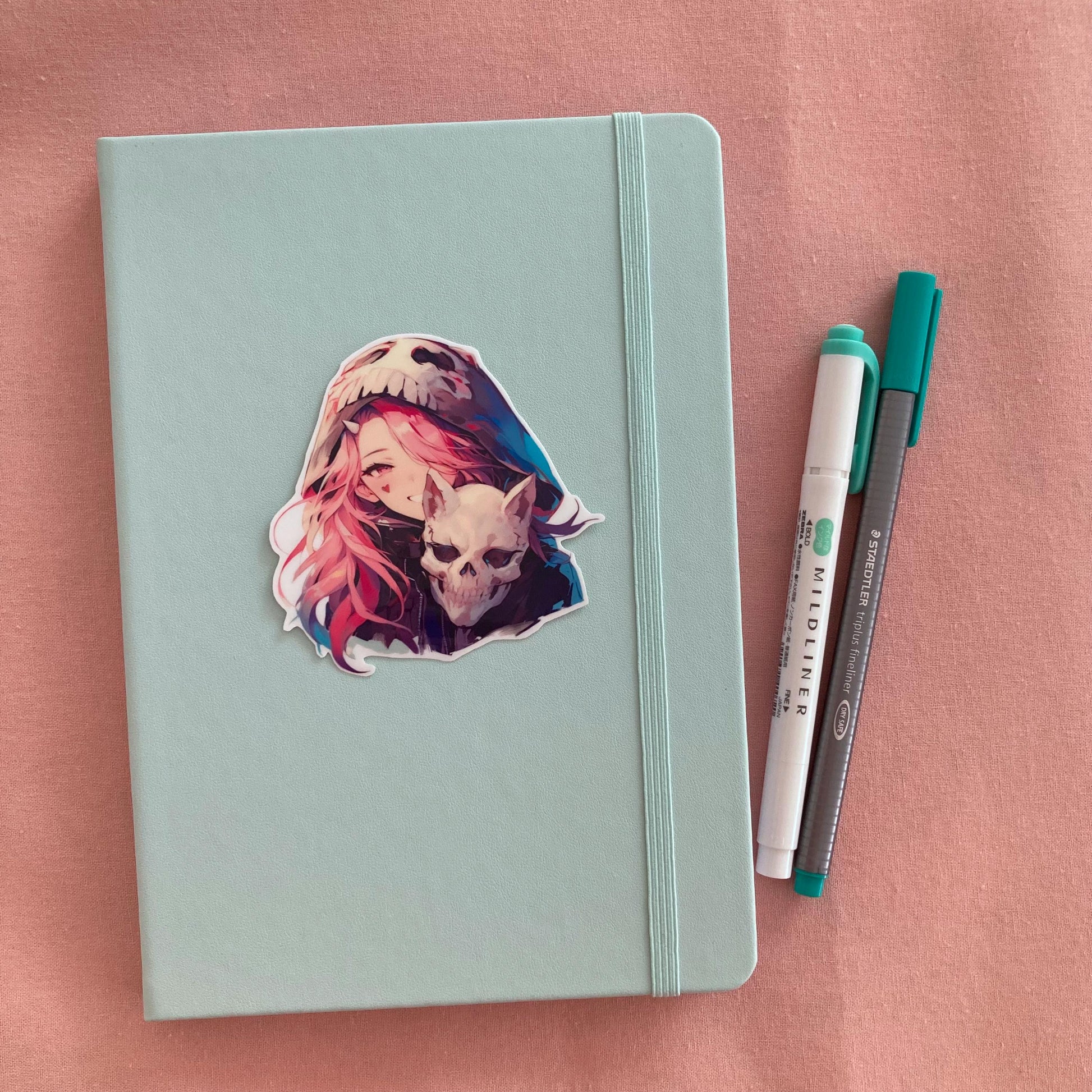 Mint green notebook with a sticker of an anime character wearing a skull-themed hood and holding a skull, placed on a pink surface. Beside the notebook are a teal pen and a white highlighter.
