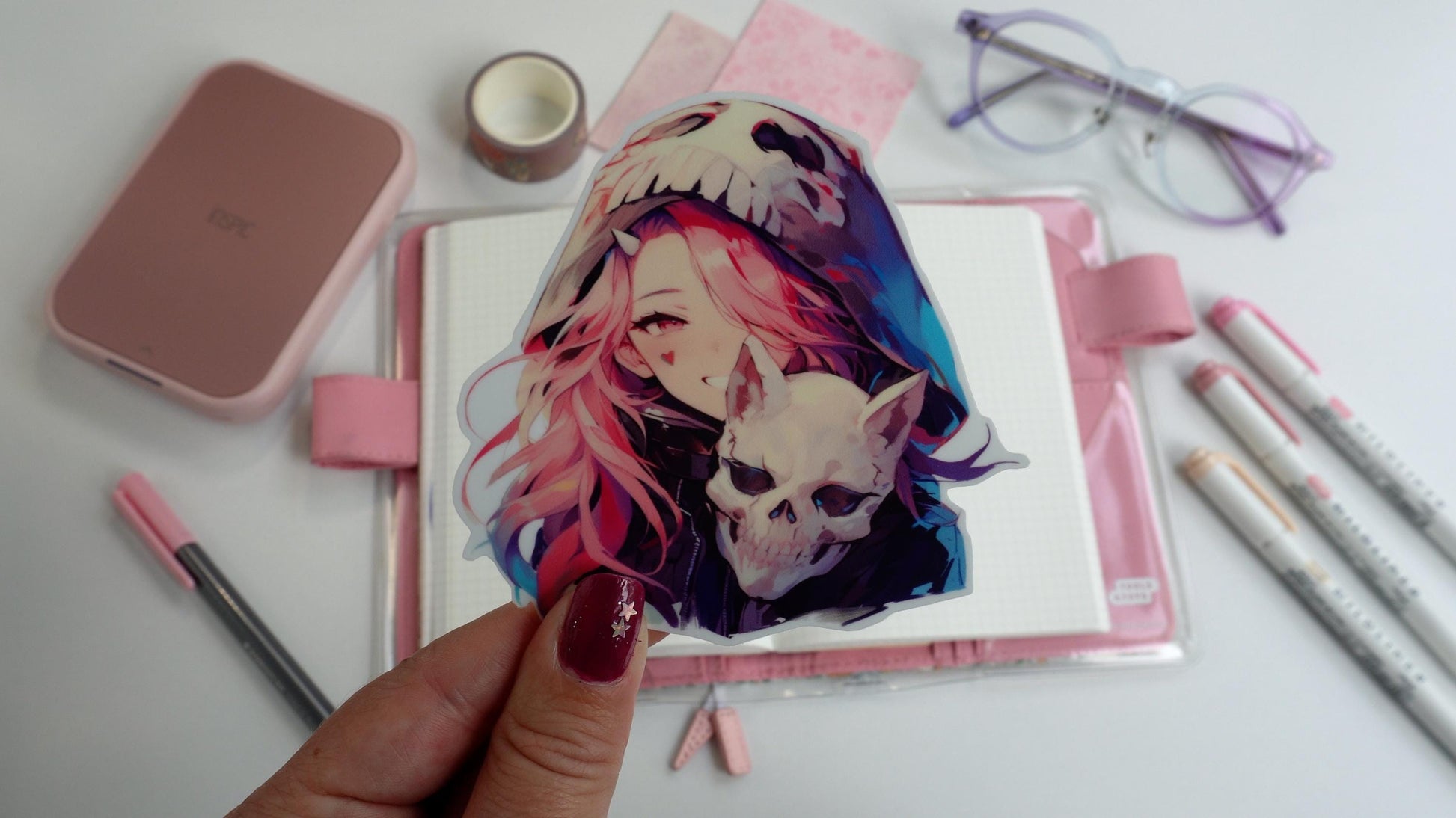 A hand holding a sticker of an anime girl with pink hair wearing a skull hood and holding a skull. The background includes a book, pink pens, purple glasses, decorative tape, and a pink device on a white surface. Nails are painted purple with stars