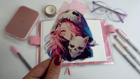 A hand holding a sticker of an anime girl with pink hair wearing a skull hood and holding a skull. The background includes a book, pink pens, purple glasses, decorative tape, and a pink device on a white surface. Nails are painted purple with stars