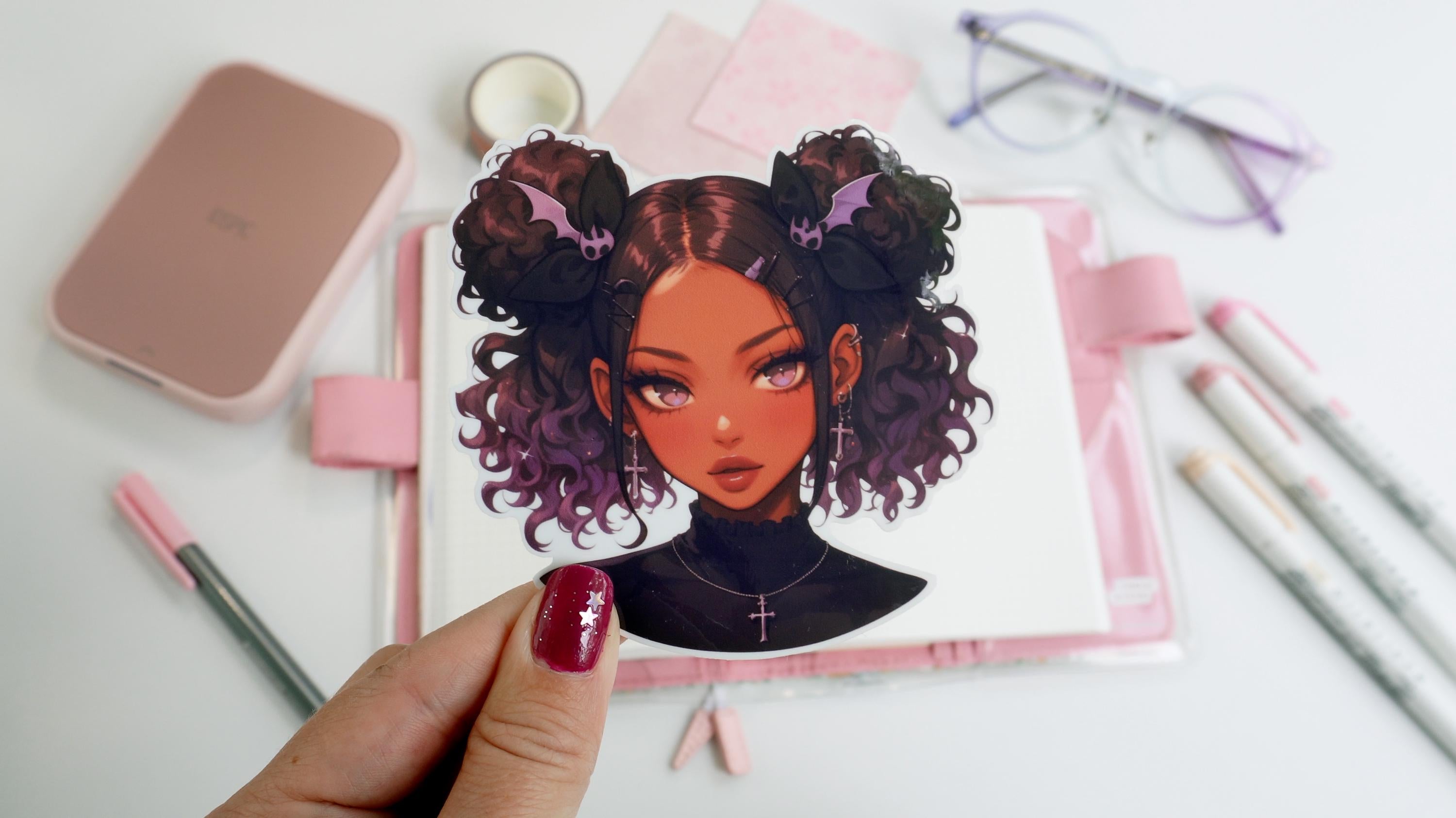 A hand with maroon nail polish and a silver star design holds a sticker of a girl with curly hair styled in buns, bat-shaped hair clips, and cross earrings. The background includes a notebook, markers, glasses, and pink paper on a white surface