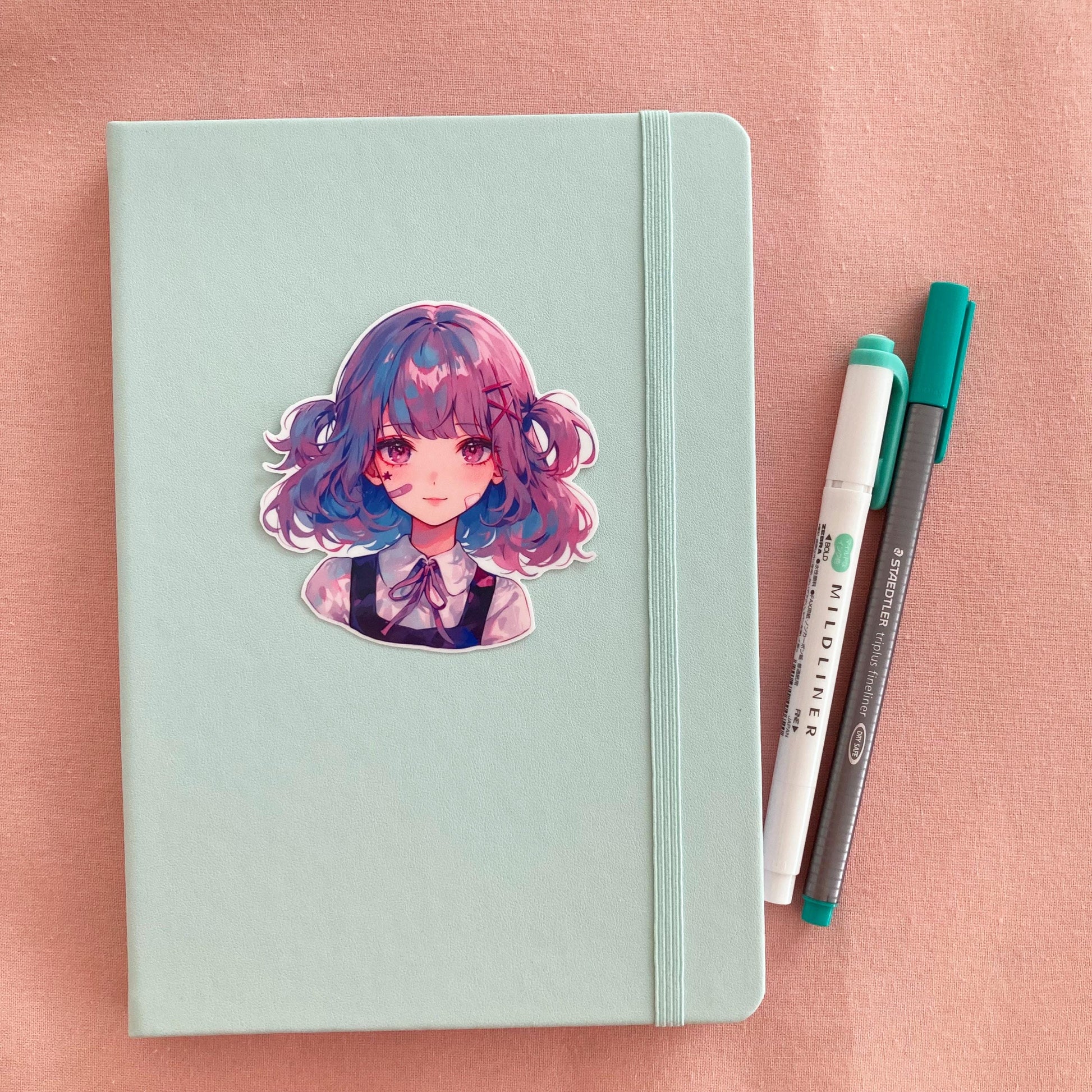 A mint green notebook with a sticker of a girl with blue and pink hair. Next to it are a white and green marker and a gray fineliner pen, all placed on a soft pink surface.