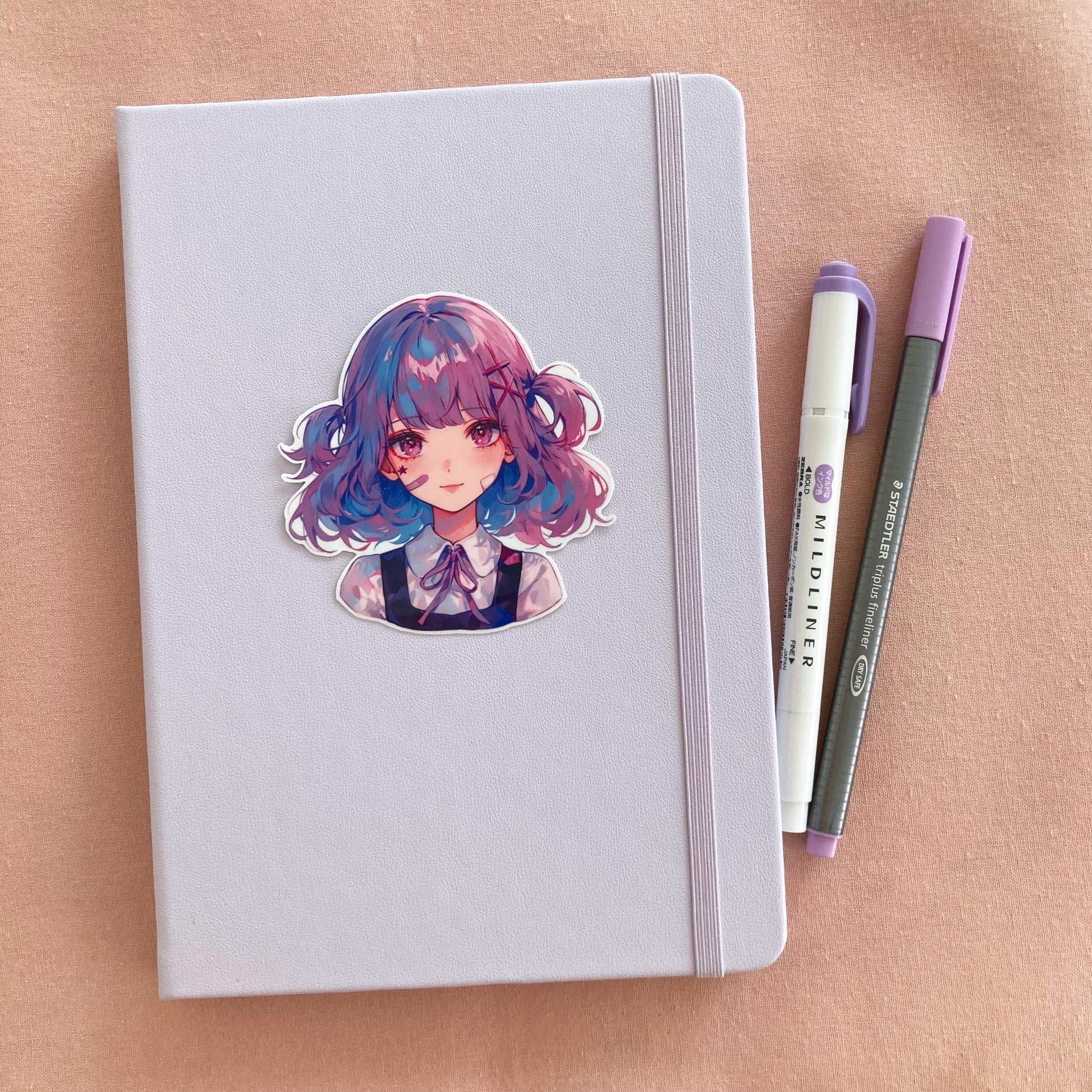 A light purple notebook with a sticker of a girl with blue and pink hair. Next to it are a white and purple marker and a gray fineliner pen, all placed on a soft pink surface.