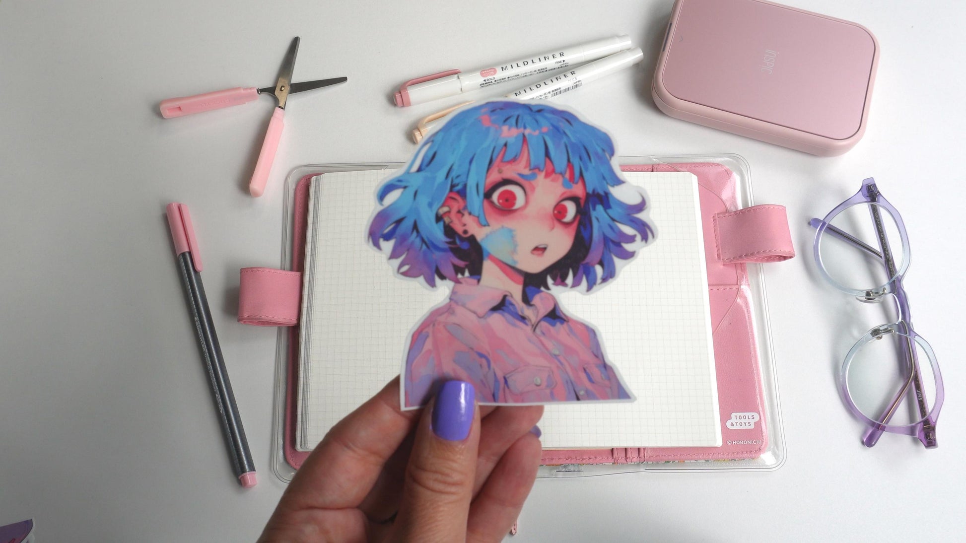 A hand with purple nails holds a colorful sticker of an anime character with blue hair and red eyes. The background includes a pink notebook, pastel pens, pink scissors, a small pink case, and purple-rimmed glasses, all arranged on a white surface