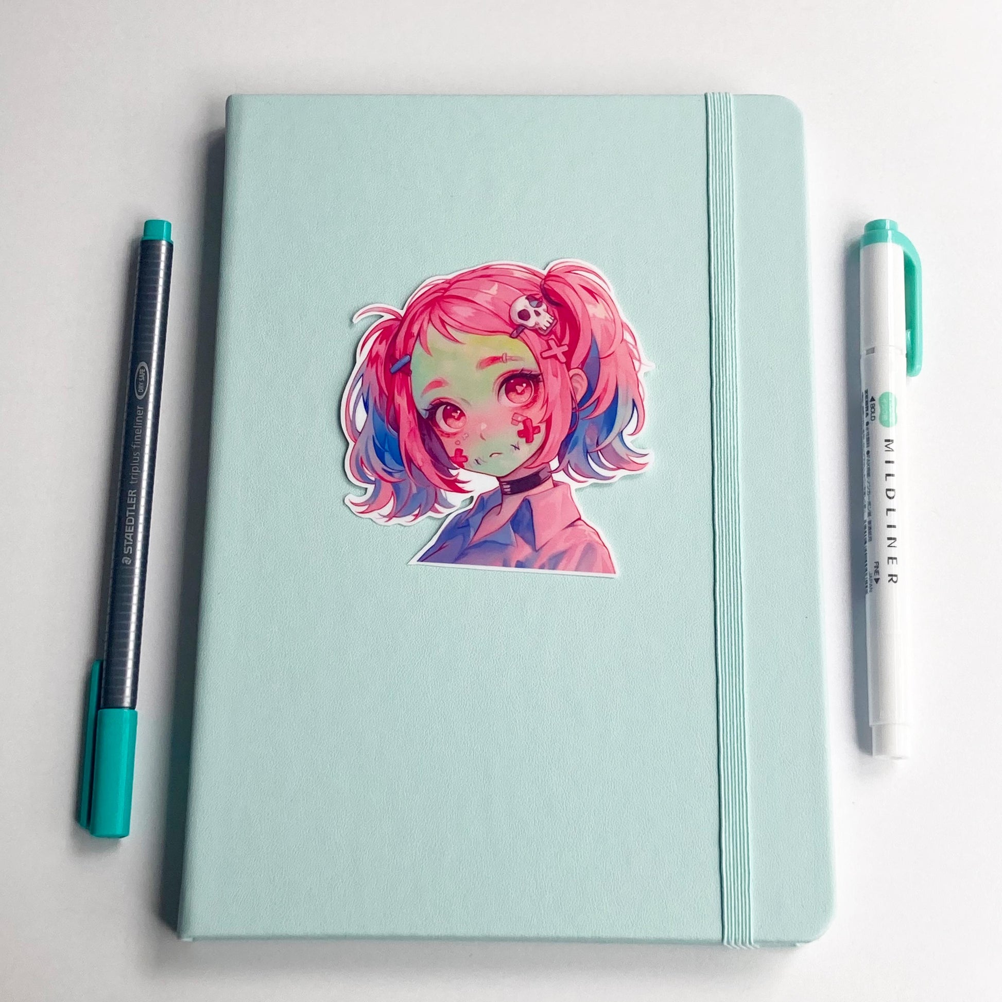 A sticker of an anime character with pink and blue hair is placed on a closed light teal notebook. A pen with a teal cap is on the left, and a teal marker is on the right.