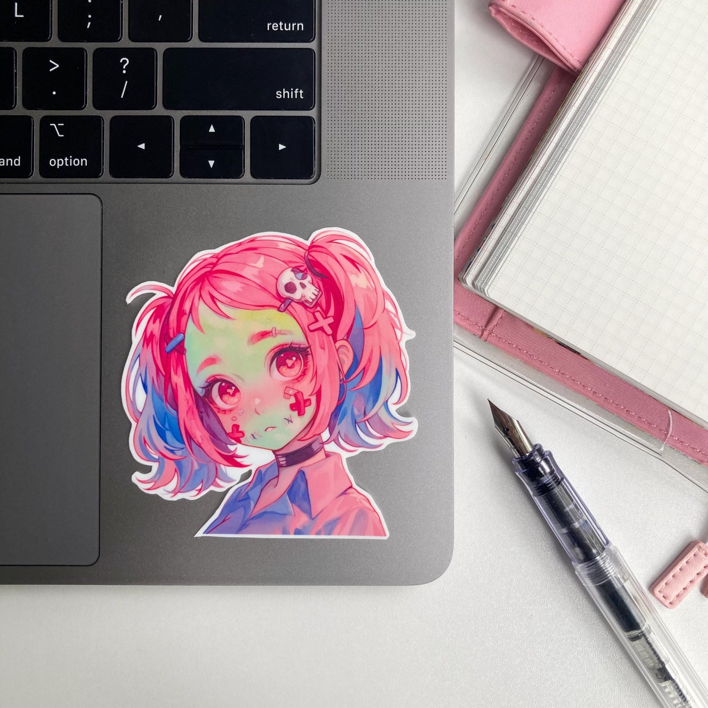 A sticker of an anime character with pink and blue hair, red eyes, and skull accessories is on a laptop. Nearby are a pink notebook and a fountain pen.