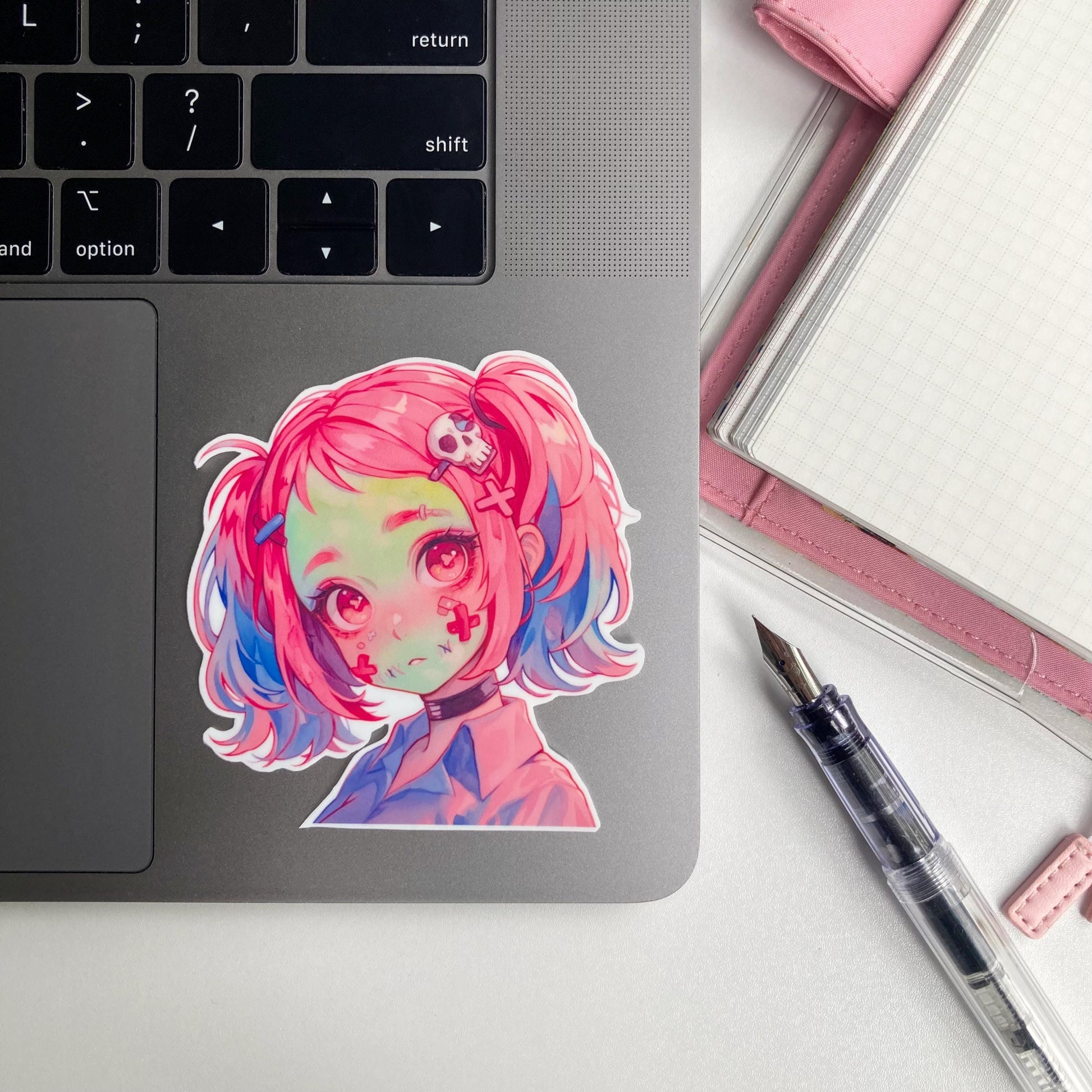 A sticker of an anime character with pink and blue hair, red eyes, and skull accessories is on a laptop. Nearby are a pink notebook and a fountain pen.