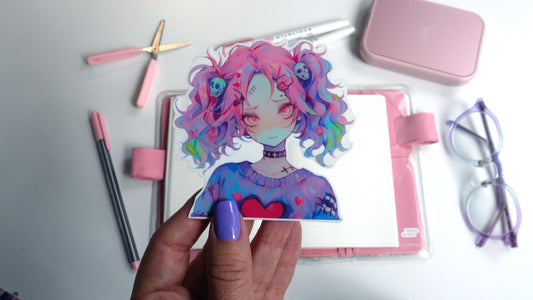 A hand with purple nails holds a sticker of an anime character with pink and multicolored hair, skull accessories, and a heart on their sweater. Background items include a pink notebook, pens, scissors, a case, and glasses on a white surface.
