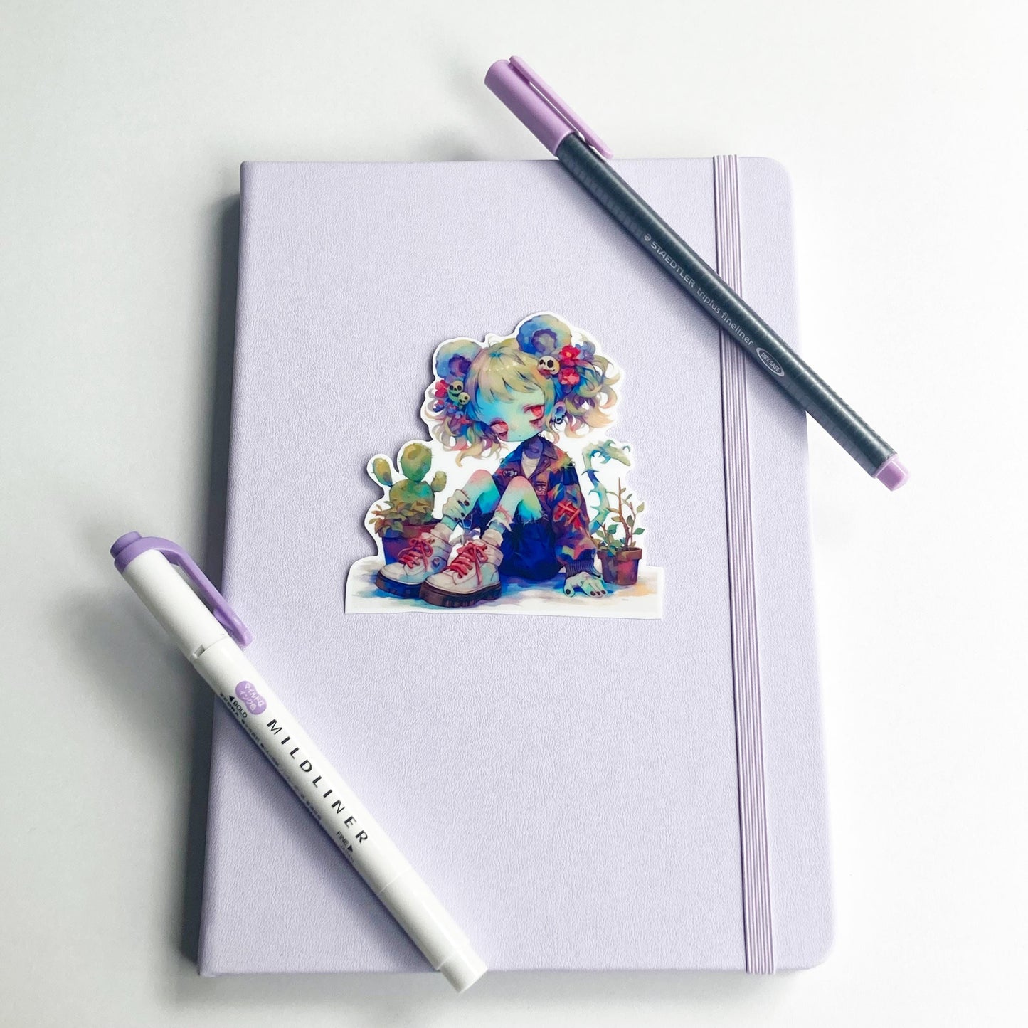 A sticker of an anime character with colorful hair, mouse ears, and potted plants is placed on a closed light purple notebook. A purple marker and a pen with a purple cap lie diagonally on the notebook.