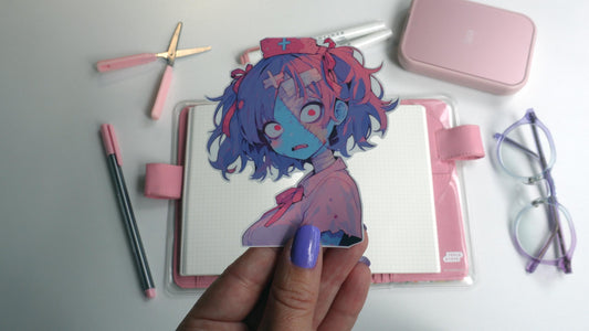A hand with purple nail polish holds a nurse zombie sticker up. The zombie has blue and pink skin, purple hair in pigtails with pink bows and wears a vintage nurses cap with a medical cross on it. The background has a journal and stationery items