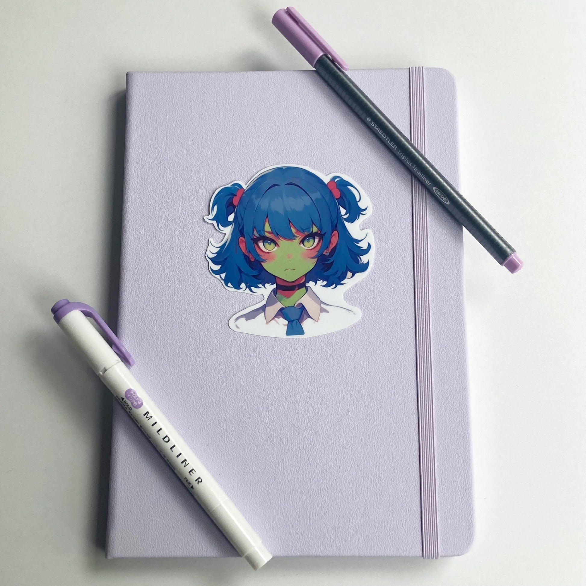 Sticker of an anime-style character with blue twin-tails, pink hair ties, green skin, and yellow eyes on a lavender notebook. The character wears a white shirt, blue tie, and black choker. A purple pen and highlighter are placed on the notebook.