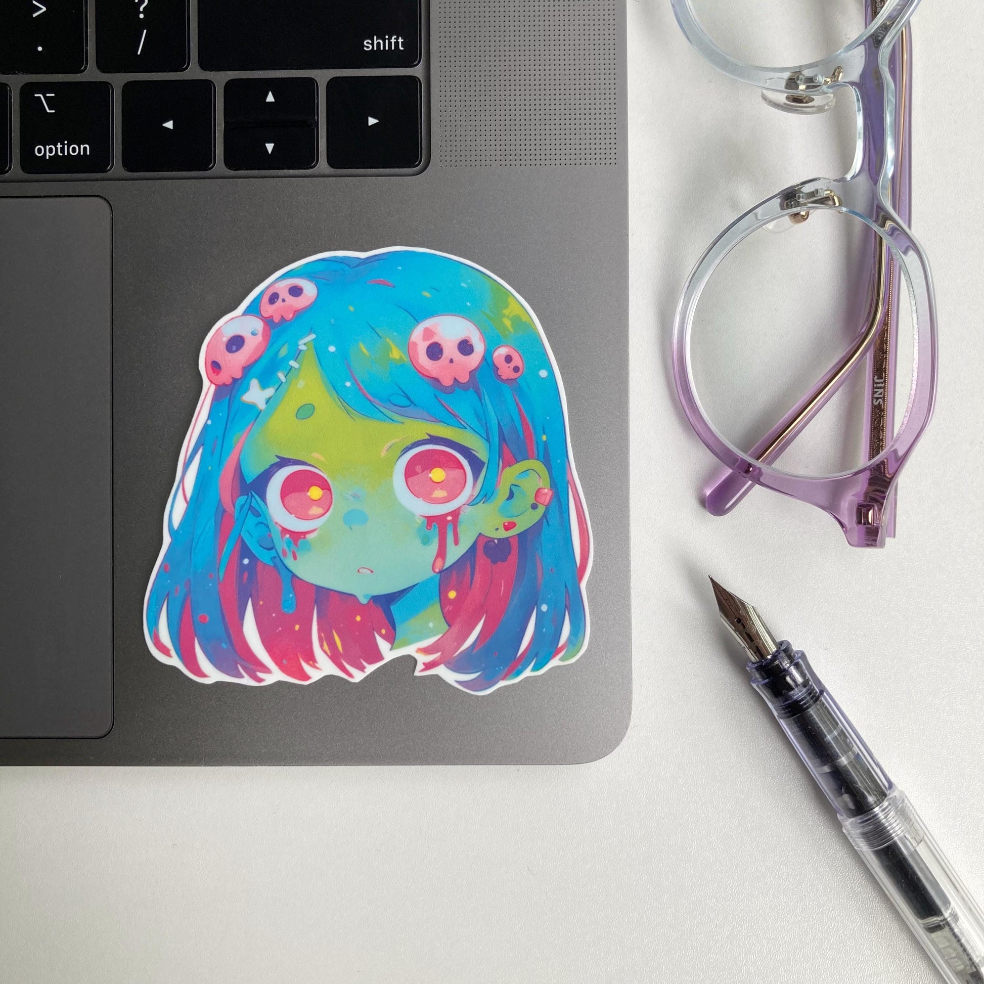 Sticker of an anime-style zombie girl with blue and pink hair, green skin, and large pink eyes. Blood drips from her eyes, and her hair is decorated with small pink skulls. The sticker is placed on a laptop keyboard corner.