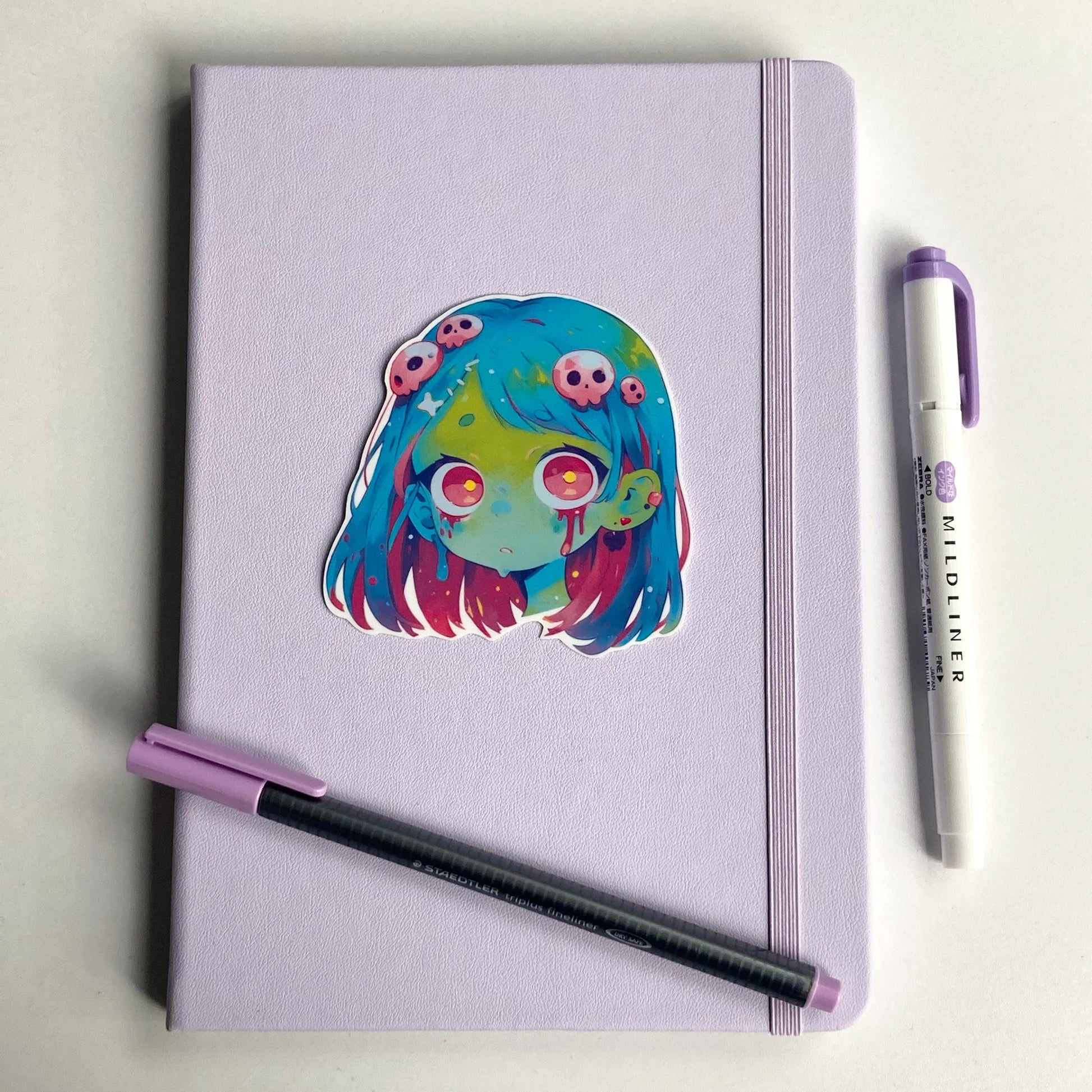 Sticker of an anime zombie girl with blue and pink hair, green skin, and large pink eyes on a lavender notebook. Blood drips from her eyes, and her hair is decorated with small pink skulls. A purple pen and matching highlighter are on the notebook.
