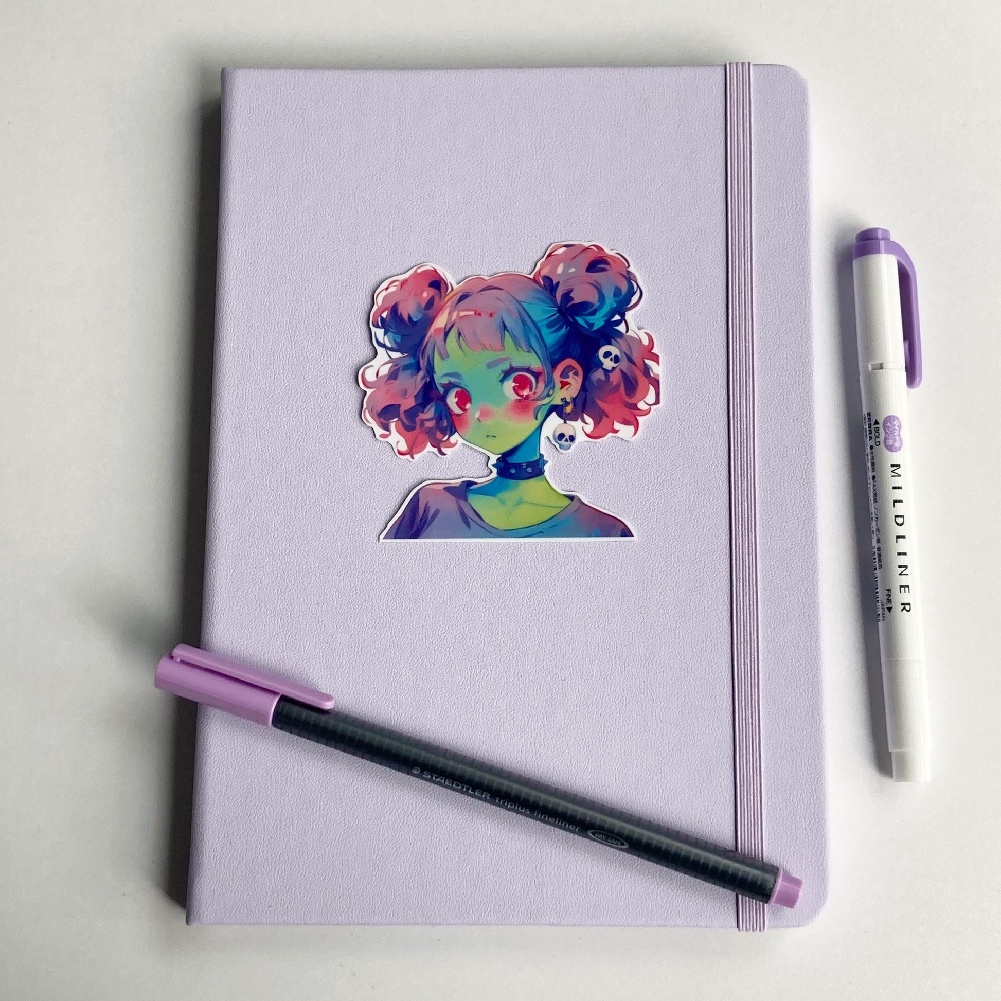 Sticker of an anime-style character with pastel pink and blue twin buns, green skin, and pink eyes on a lavender notebook. The character wears a black choker and skull earrings. A purple pen and matching highlighter are placed on the notebook.