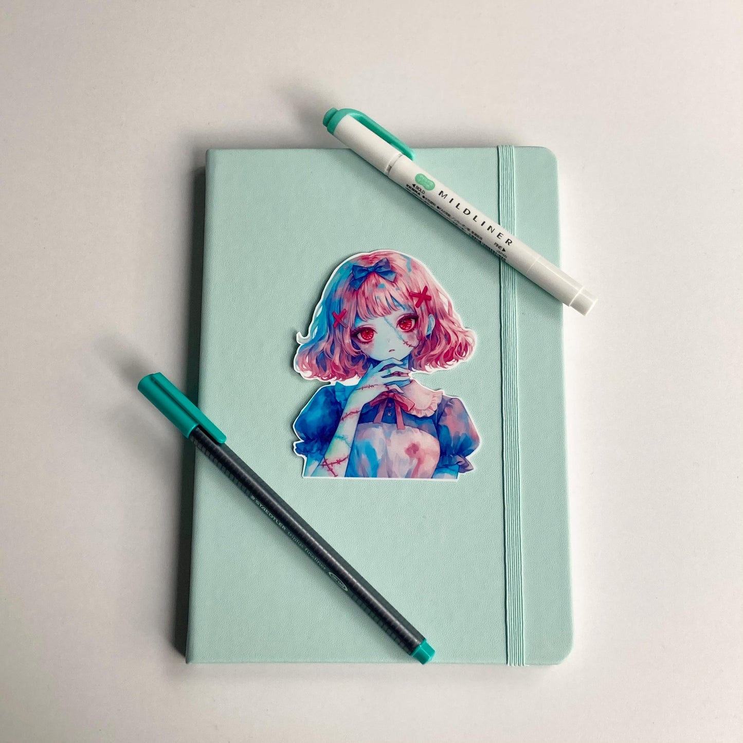 A colorful anime-style sticker of a girl with pink hair and red eyes is placed on a mint green notebook. A black pen with a teal cap and a matching teal highlighter are positioned beside the notebook on a white surface.