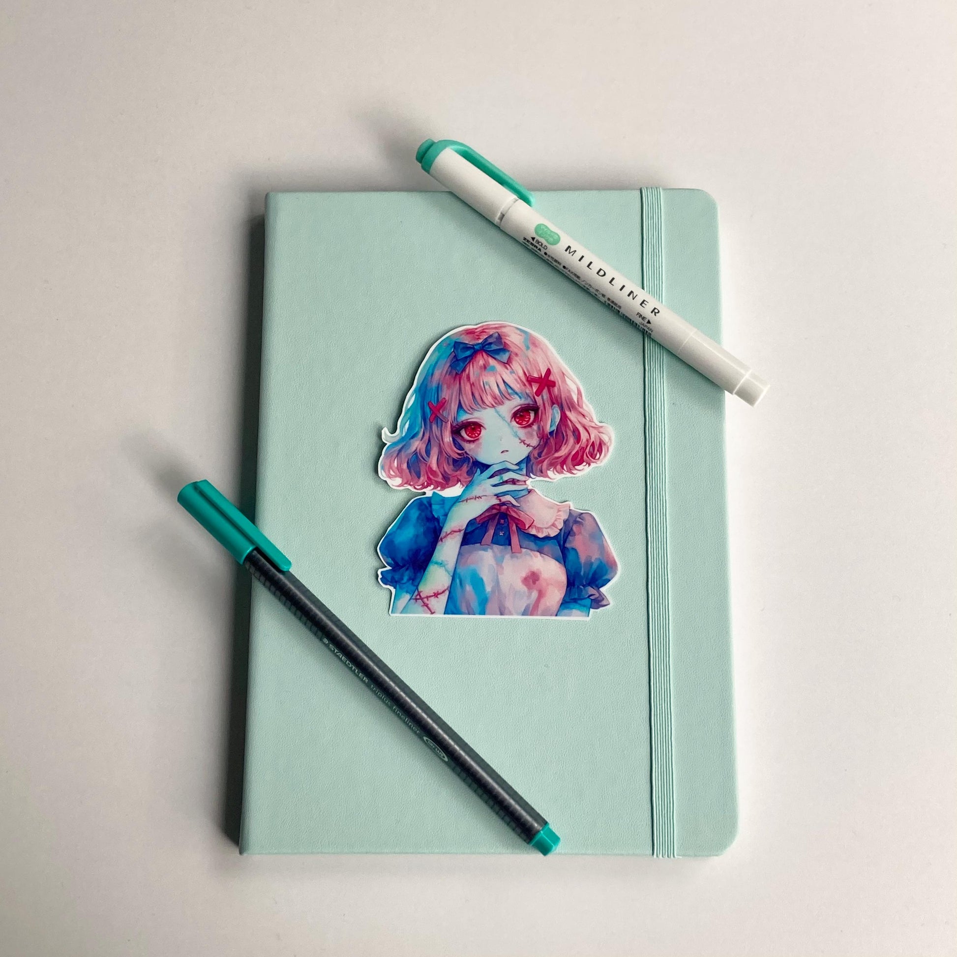 A colorful anime-style sticker of a girl with pink hair and red eyes is placed on a mint green notebook. A black pen with a teal cap and a matching teal highlighter are positioned beside the notebook on a white surface.