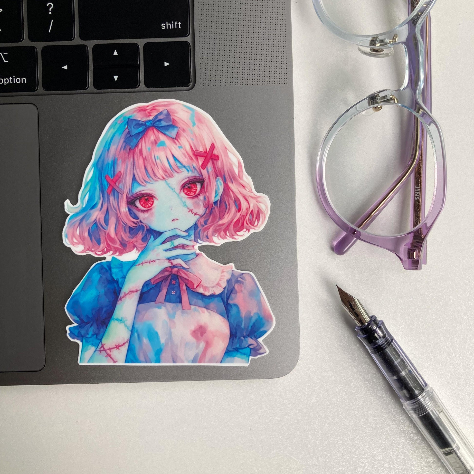 A colorful anime-style sticker of a girl with pink hair, red eyes, and stitches on her skin is placed on a laptop&#39;s gray keyboard. Nearby are purple-rimmed glasses and a fountain pen on a white surface.