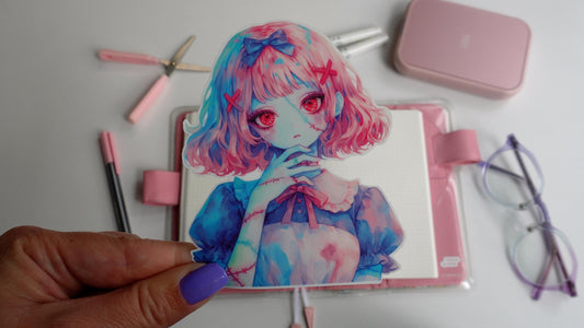 A hand with purple nails holds a colorful sticker of an anime-style girl with pink hair, red eyes, and stitches on her skin. The background features a pink notebook, pen, scissors, and glasses on a white surface.