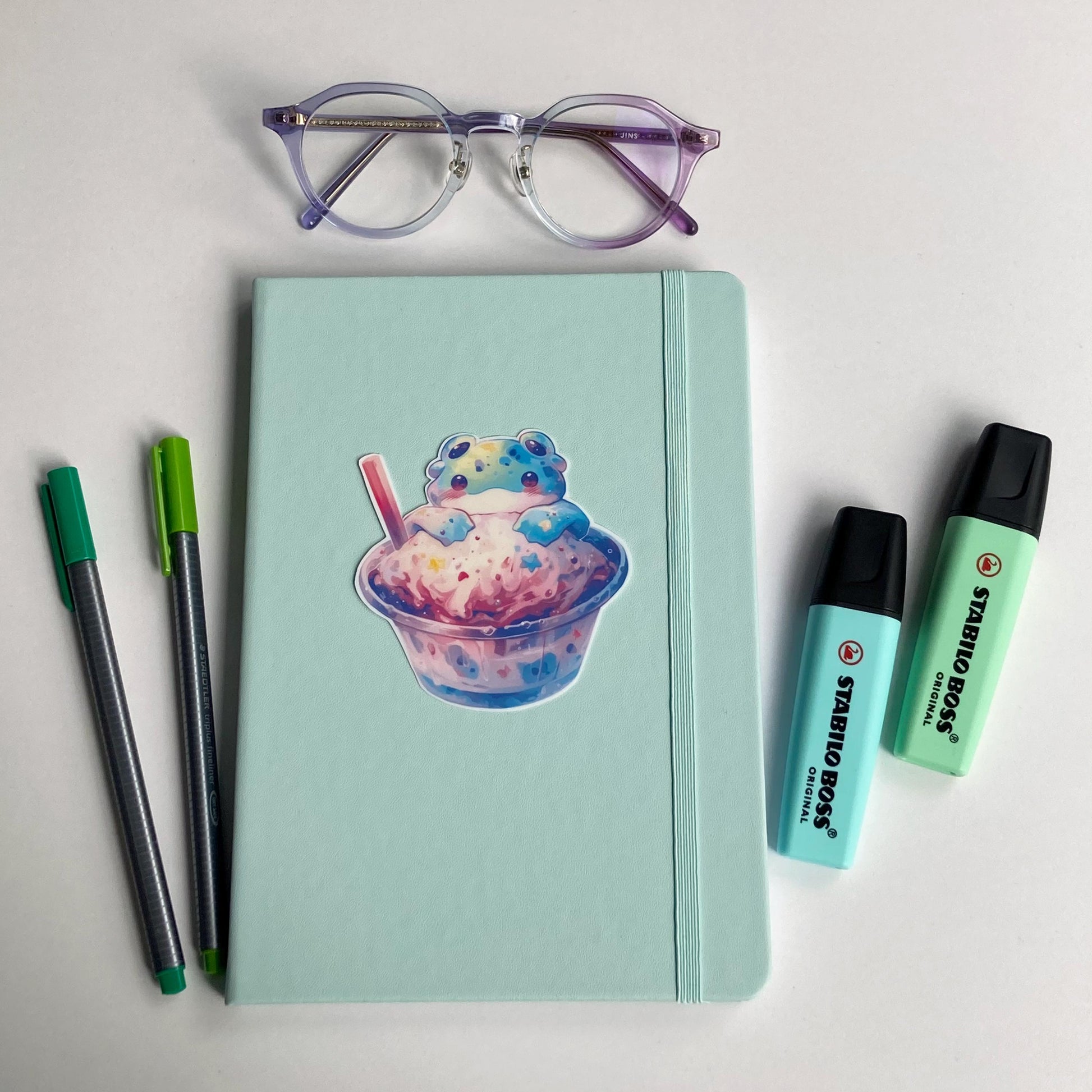 A mint green notebook with a sticker of an alien frog in a cup of pastel ice cream is surrounded by purple-rimmed glasses, two green pens, and two mint-colored highlighters. The items are arranged on a white surface.