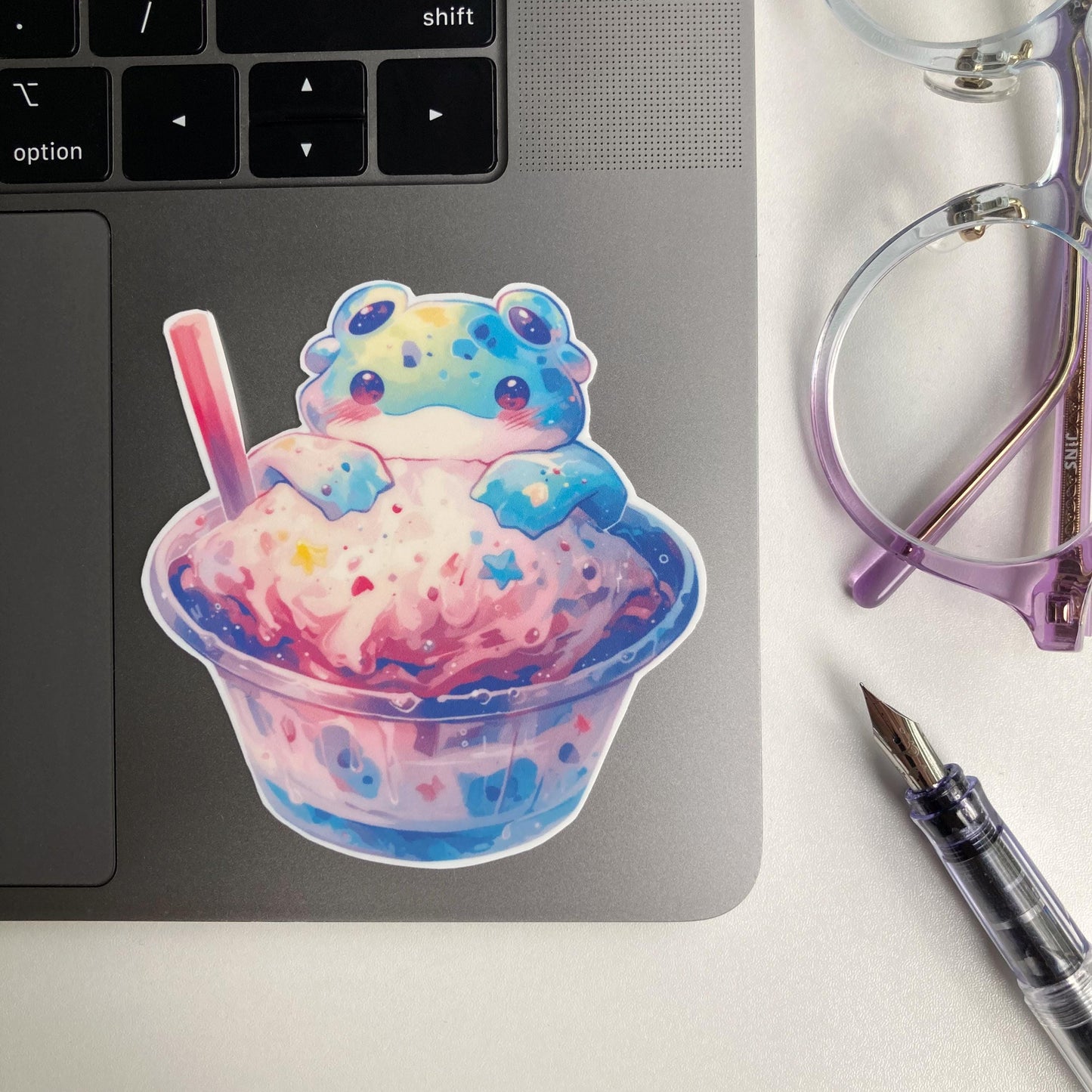 A colorful sticker of an alien frog sitting in a cup of pastel pink and blue ice cream is placed on a laptop. Nearby are a pair of purple-rimmed glasses and a clear fountain pen.