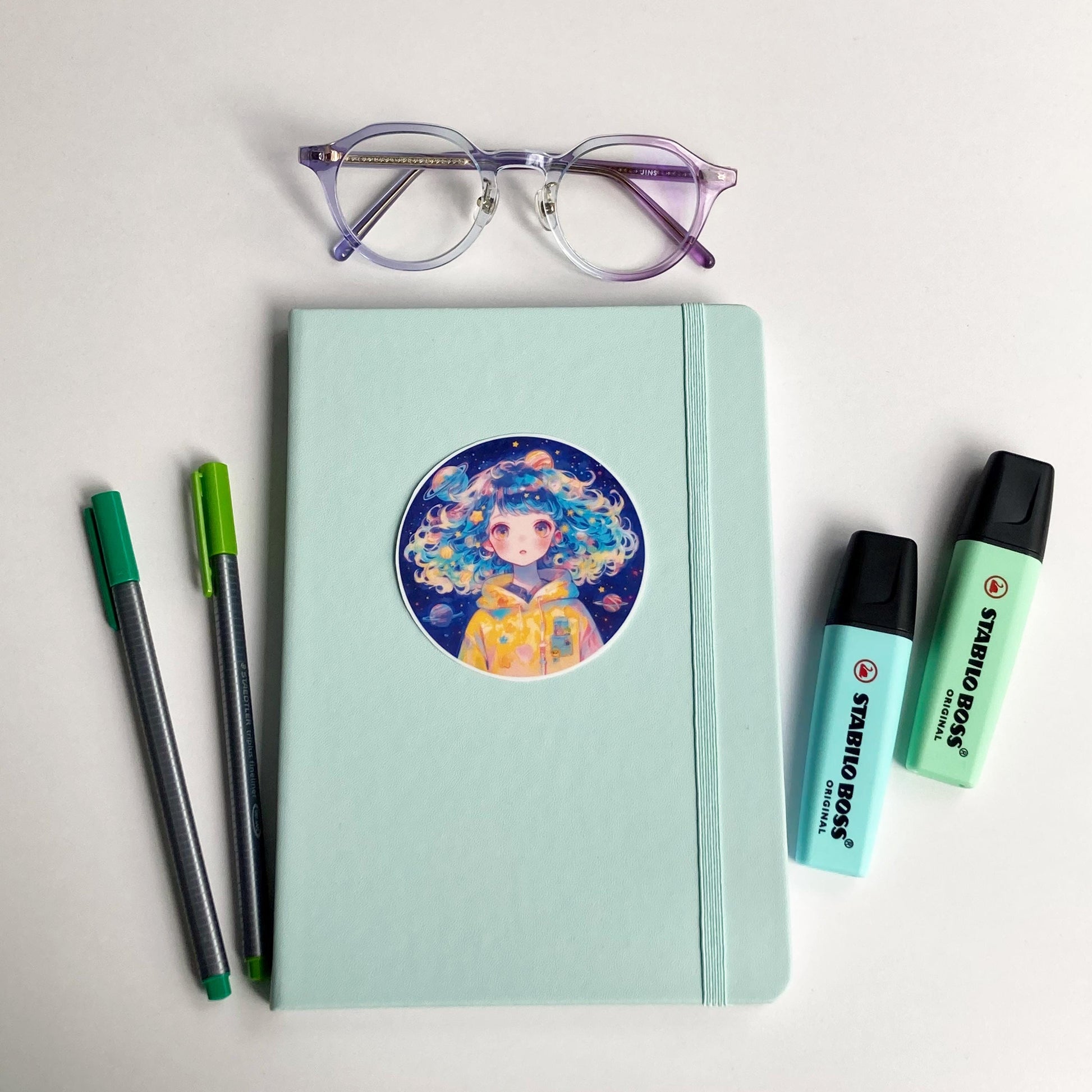 A mint green notebook with a circular sticker of a person with colorful, curly hair wearing a yellow space-themed hoodie is surrounded by purple-rimmed glasses, two green pens, and two mint-colored highlighters.