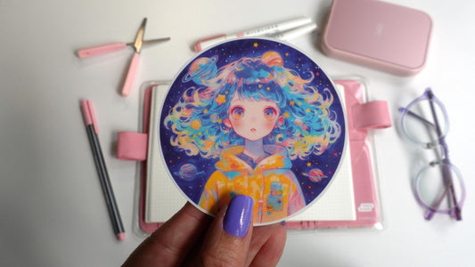 A hand with lavender nails holds a circular sticker of a person with curly blue hair wearing a yellow hoodie with space-themed designs. The background includes pink and purple stationery, a notebook, and a pair of clear glasses with purple accents