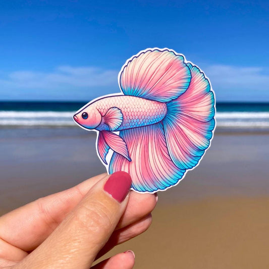 A hand with pink nails holds a sticker of a colorful betta fish with flowing pink and blue fins. The background shows a sandy beach and a bright blue ocean under a clear sky. The fish&#39;s intricate details stand out against the serene beach scene.