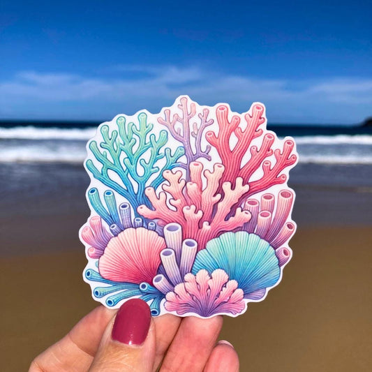 A hand holds a colorful coral reef sticker featuring various coral types in pink, blue, purple, and green. The background shows a sandy beach with waves and a clear blue sky, highlighting the intricate reef details against the coastal scene