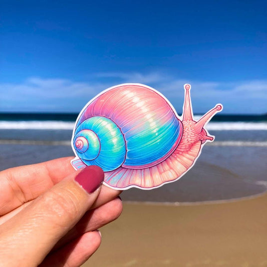 A hand holds a colorful snail sticker with a vibrant shell in shades of pink, blue, and purple. The background shows a sandy beach with waves and a clear blue sky, highlighting the snail&#39;s intricate details against the serene beach scene