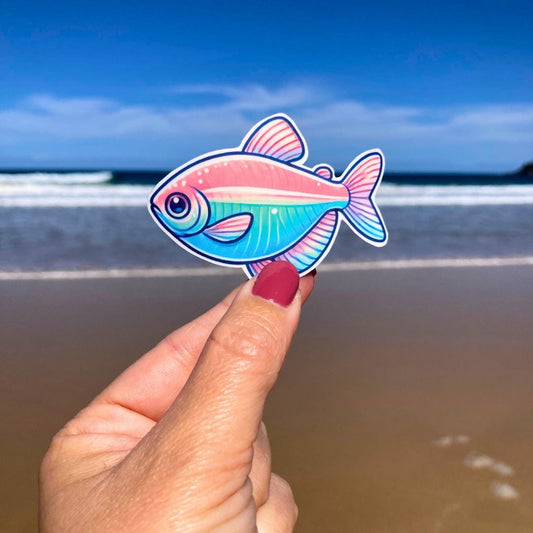 A hand with pink nails holds a colorful fish sticker featuring vibrant shades of pink, blue, and teal. The background shows a sandy beach with the ocean and clear blue sky, highlighting the contrast between the fish design and the coastal scenery.