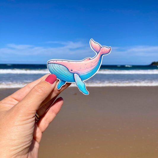 A hand with pink nails holds a colorful whale sticker with shades of pink and blue. The background shows a sandy beach with waves and a clear blue sky, highlighting the whale&#39;s playful design against the serene coastal setting.