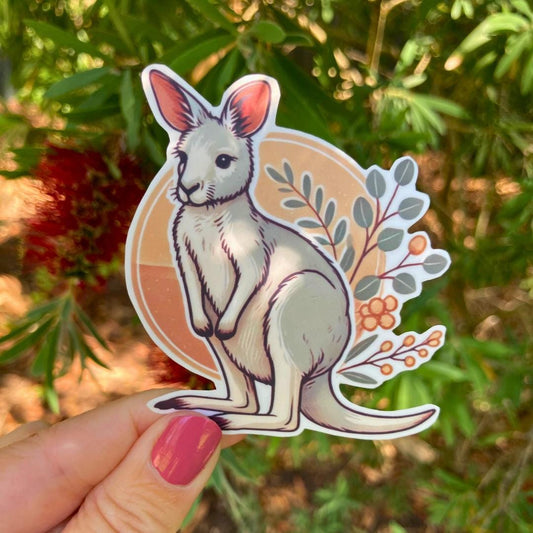 A hand with pink nail polish holds a sticker of a cartoon kangaroo with a tan circle and leafy branches in the background. The kangaroo is white with soft shading. Behind it, blurred green leaves and red bottlebrush flowers are visible.