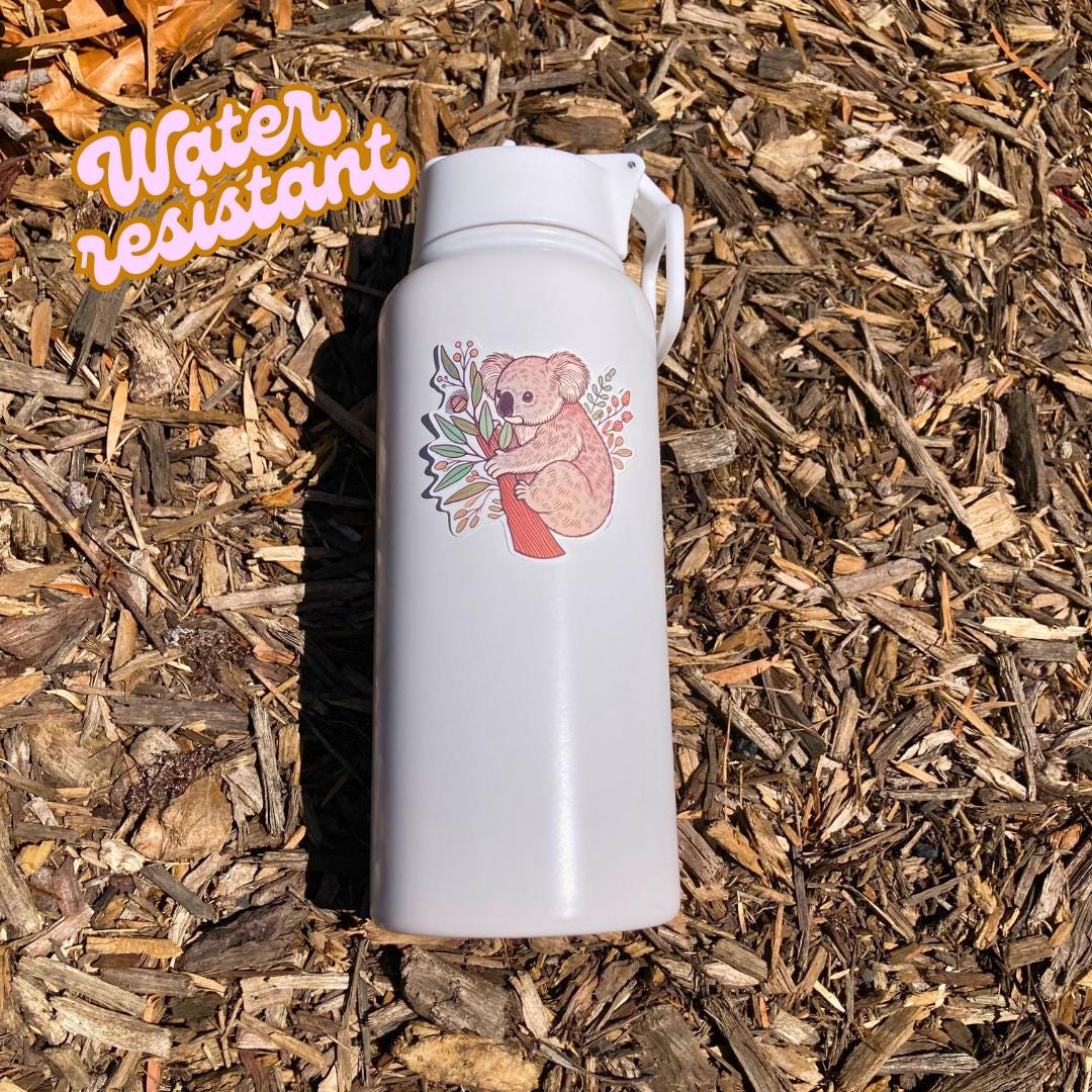 A white water bottle with a sticker of a koala hugging a tree trunk with eucalyptus leaves and branches lies on wood chips in bright sunlight. The koala has soft brown fur. An orange &quot;Water resistant&quot; label is in the top left corner.