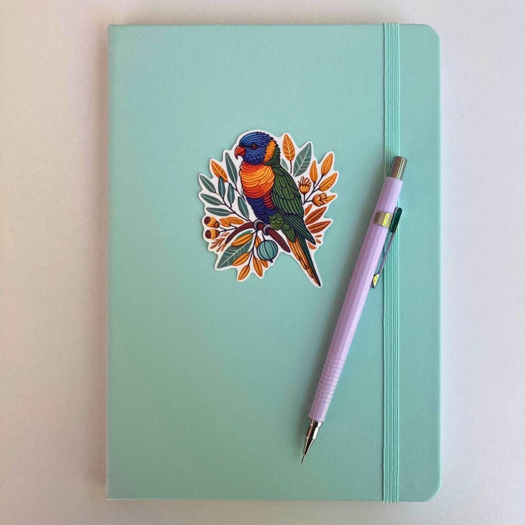 A light turquoise notebook with an elastic strap features a sticker of a colorful rainbow lorikeet perched on a branch with orange and green leaves. A lavender mechanical pencil is placed diagonally on the notebook, which rests on a white surface.